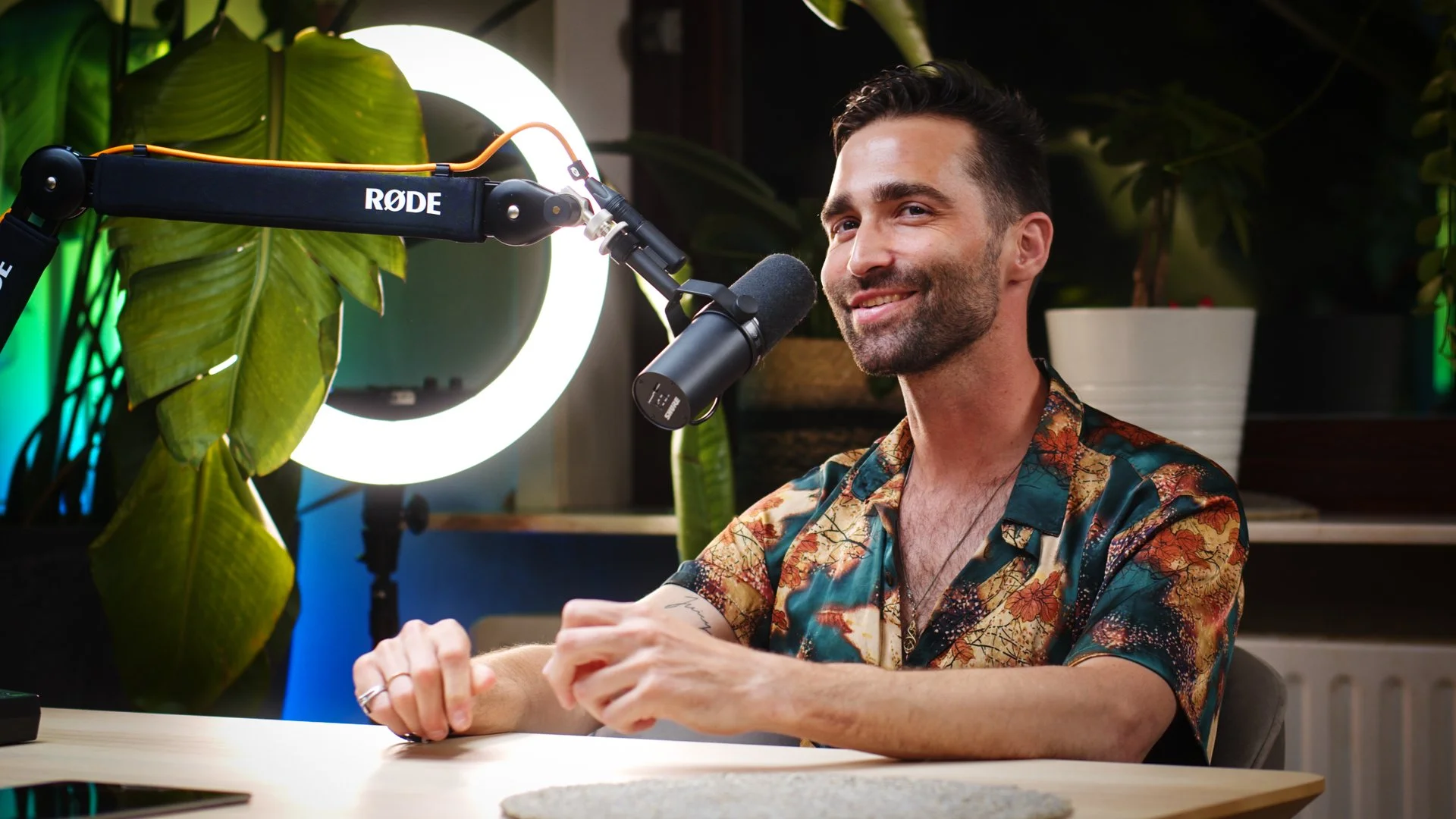 A man sitting at a desk with a microphone, smiling, in front of a ring light and green plants.