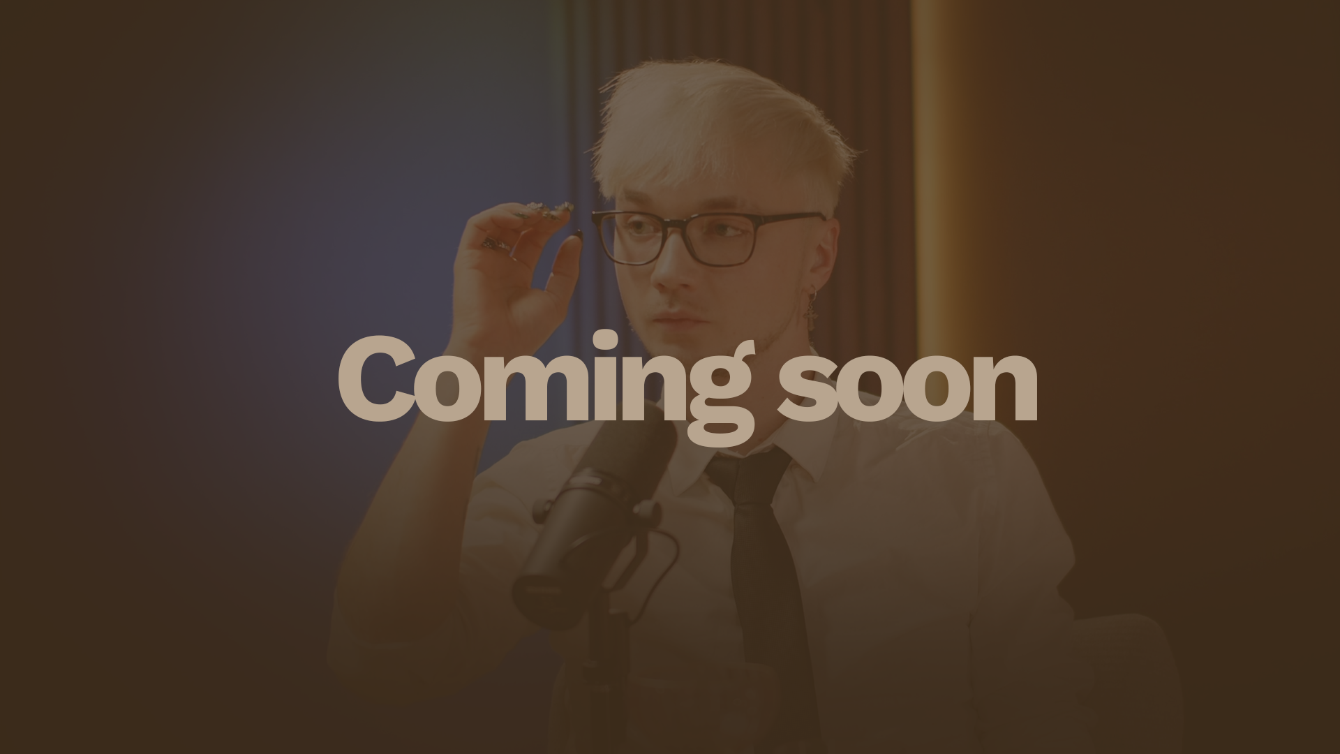 A young man with glasses, blonde hair, and earrings adjusting his glasses while sitting at a table with a microphone, a glass, and a pink bowl in front of him, in a brightly lit room with colorful curtains.