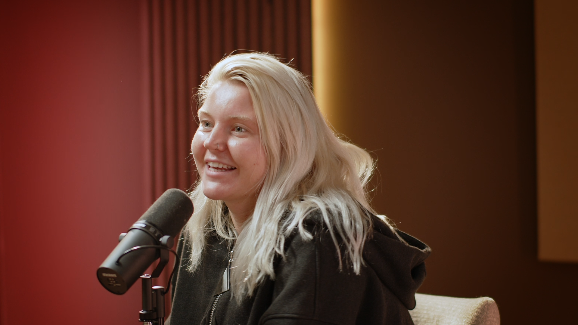 A woman with long blonde hair and a nose ring speaking into a microphone in a recording studio.