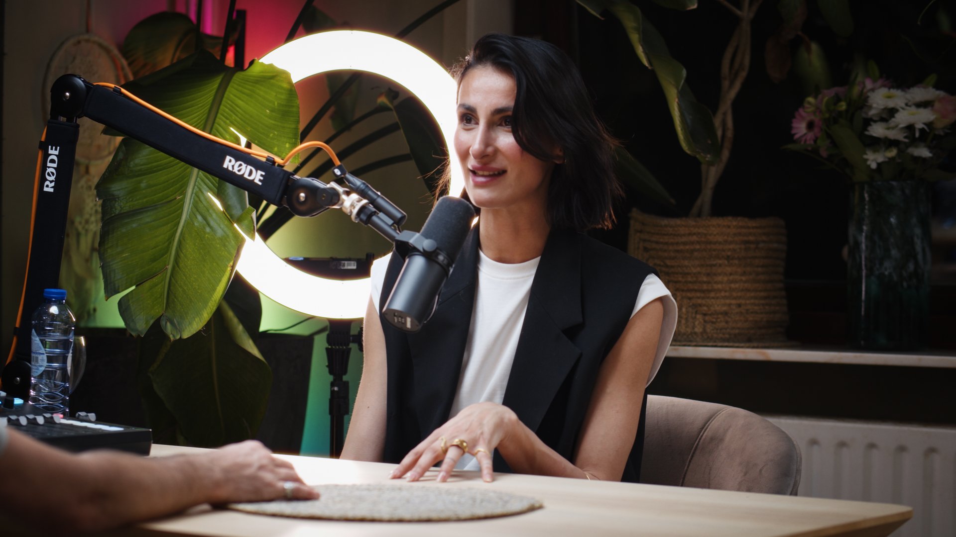 Woman speaking into a microphone during a recording session with a ring light, large green leaf, and a water bottle on the table.