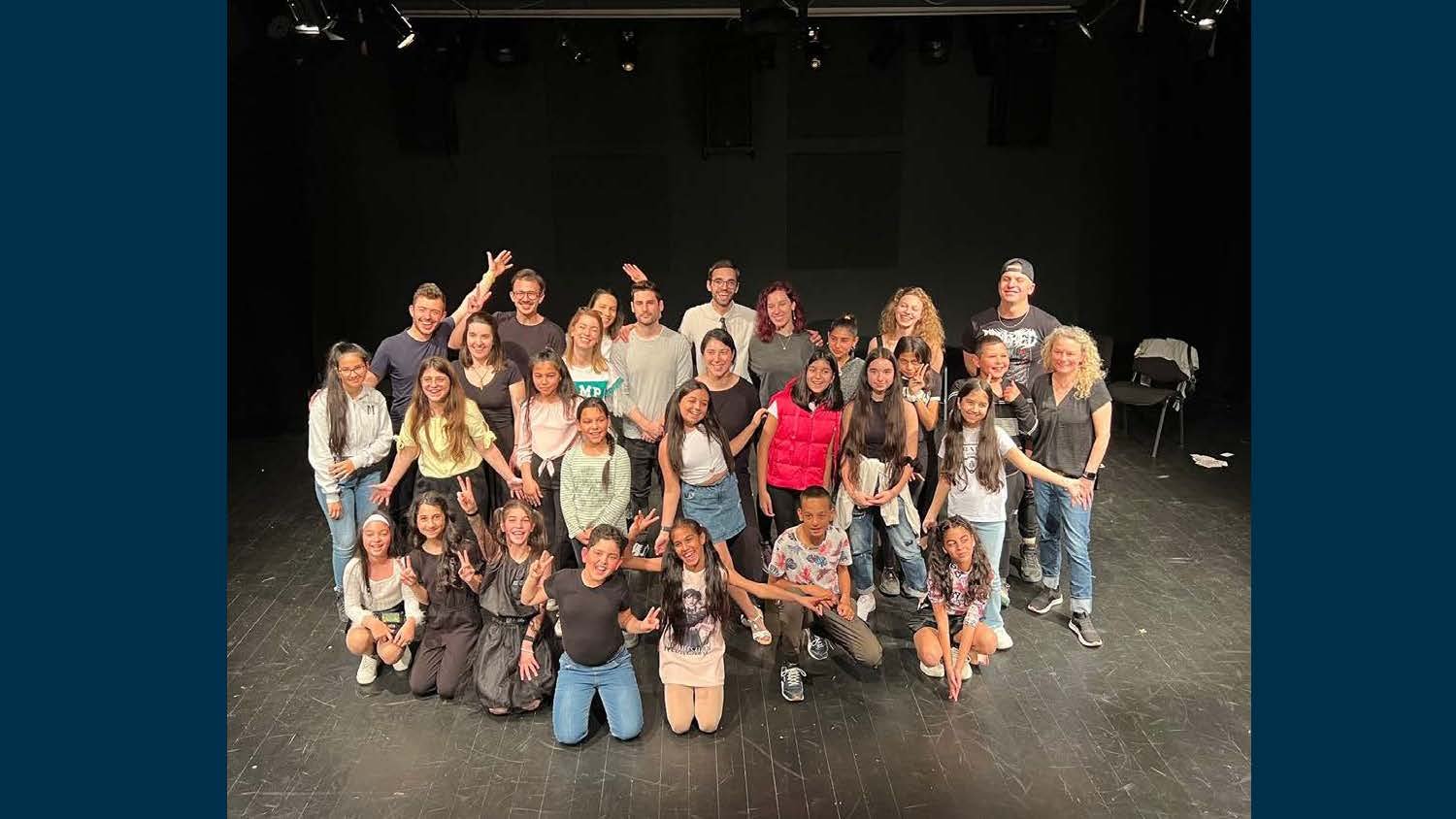Group of children and adults on a stage, smiling for a photo, in a theater or auditorium setting.