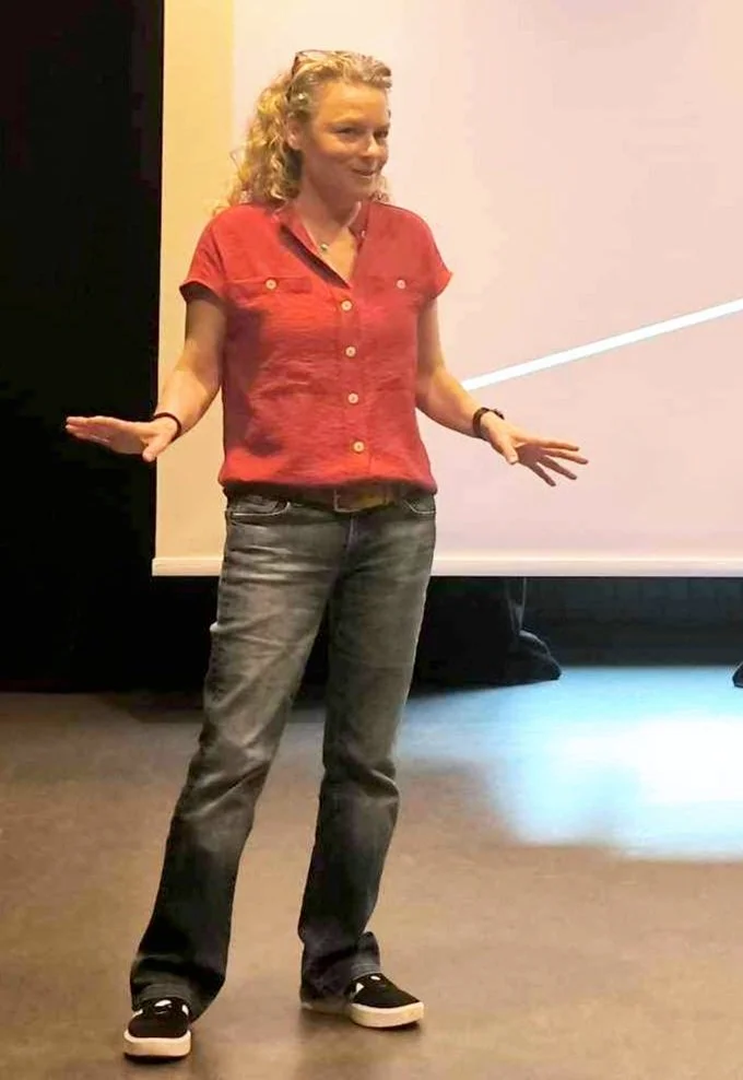 A woman with curly blonde hair wearing a red short-sleeved shirt, gray jeans, and black sneakers, standing with her hands out to the sides and smiling in a room with a white wall and projection screen.