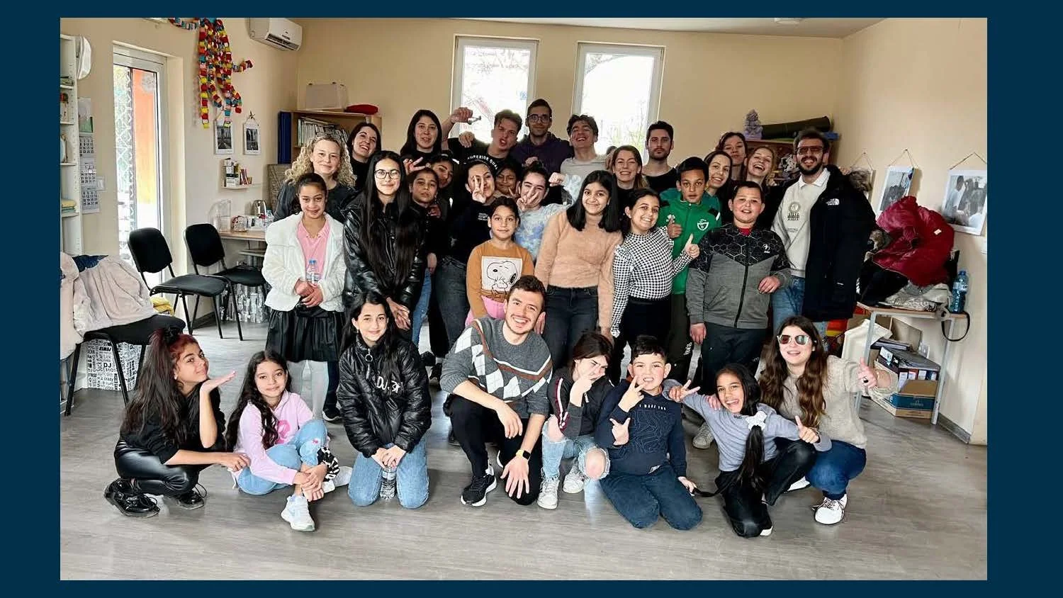 A large group of diverse children and adults posing together in a room with yellow walls and windows, some smiling, making gestures, and showing thumbs up.