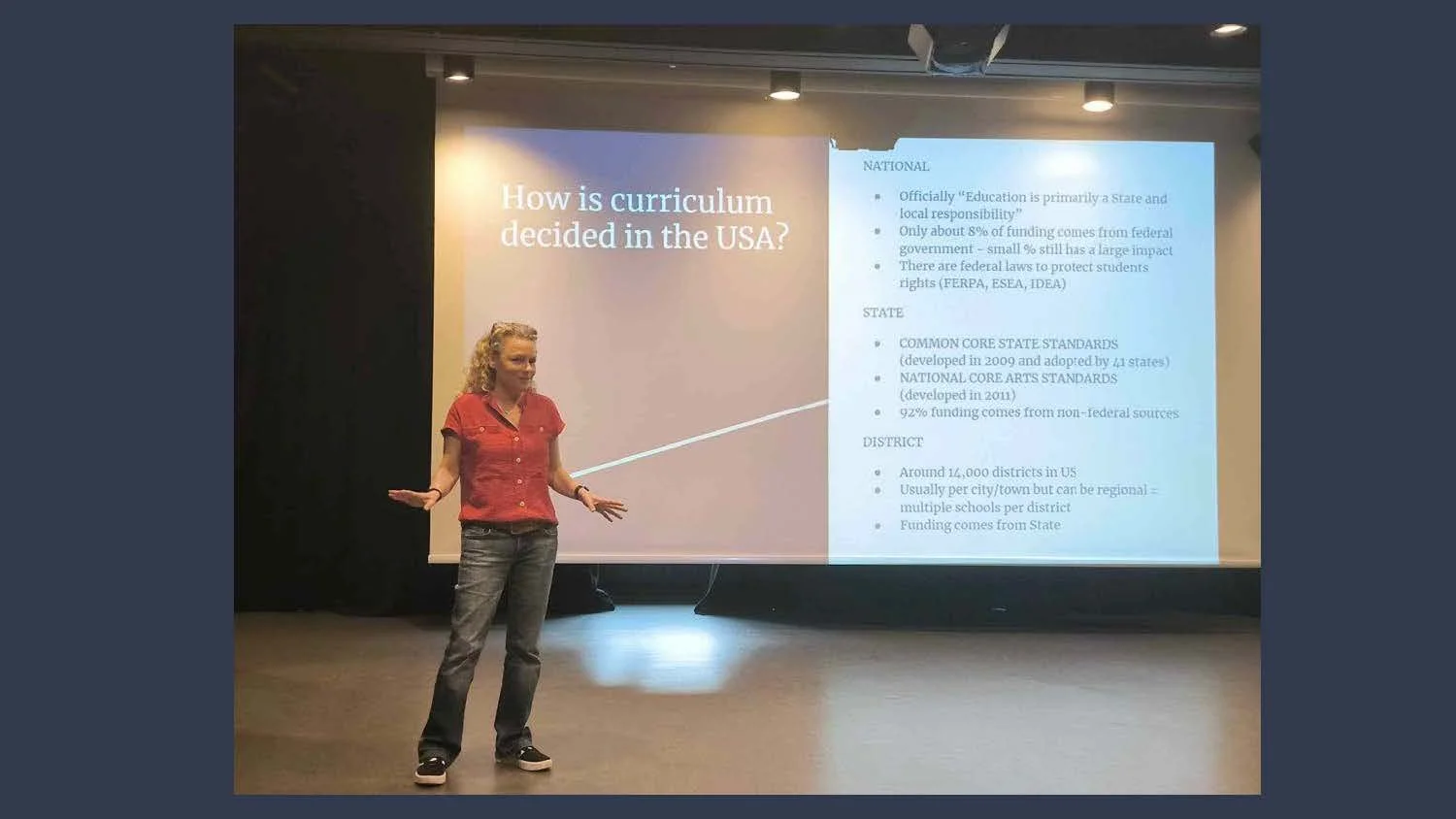 A woman in casual clothes giving a presentation on curriculum decisions in the USA, standing next to a large screen with a slide titled "How is curriculum decided in the USA?" which contains bullet points about national, state, and district levels.