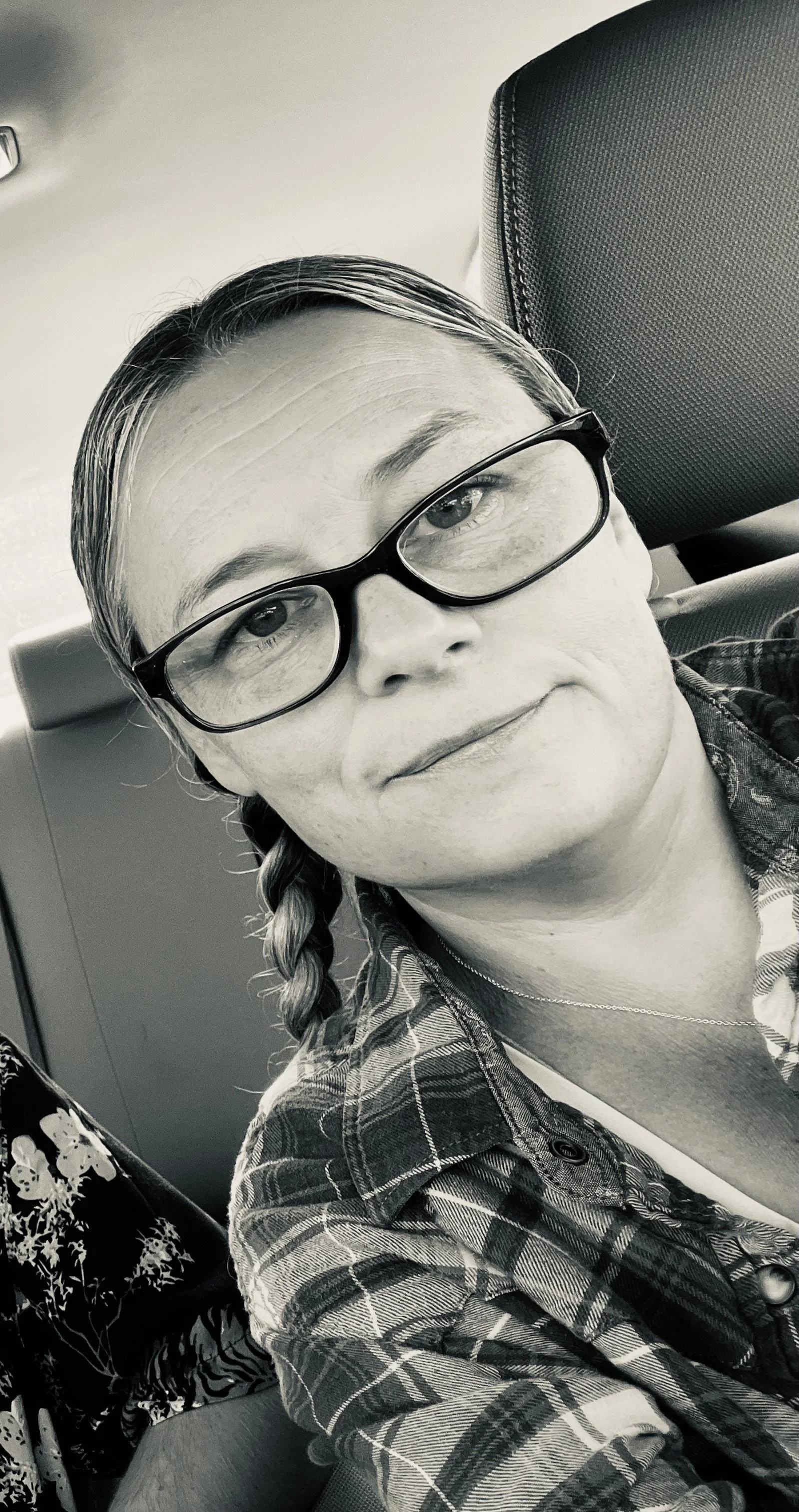A black and white photo of a woman with glasses, braided hair, wearing a plaid shirt, sitting in a car.