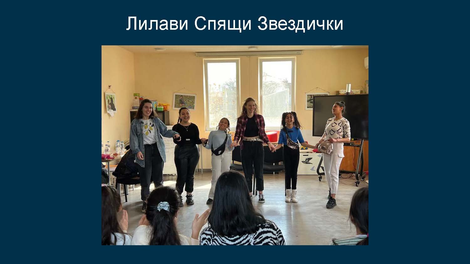 Group of people dancing and holding hands in a classroom during a performance with children watching in the foreground.