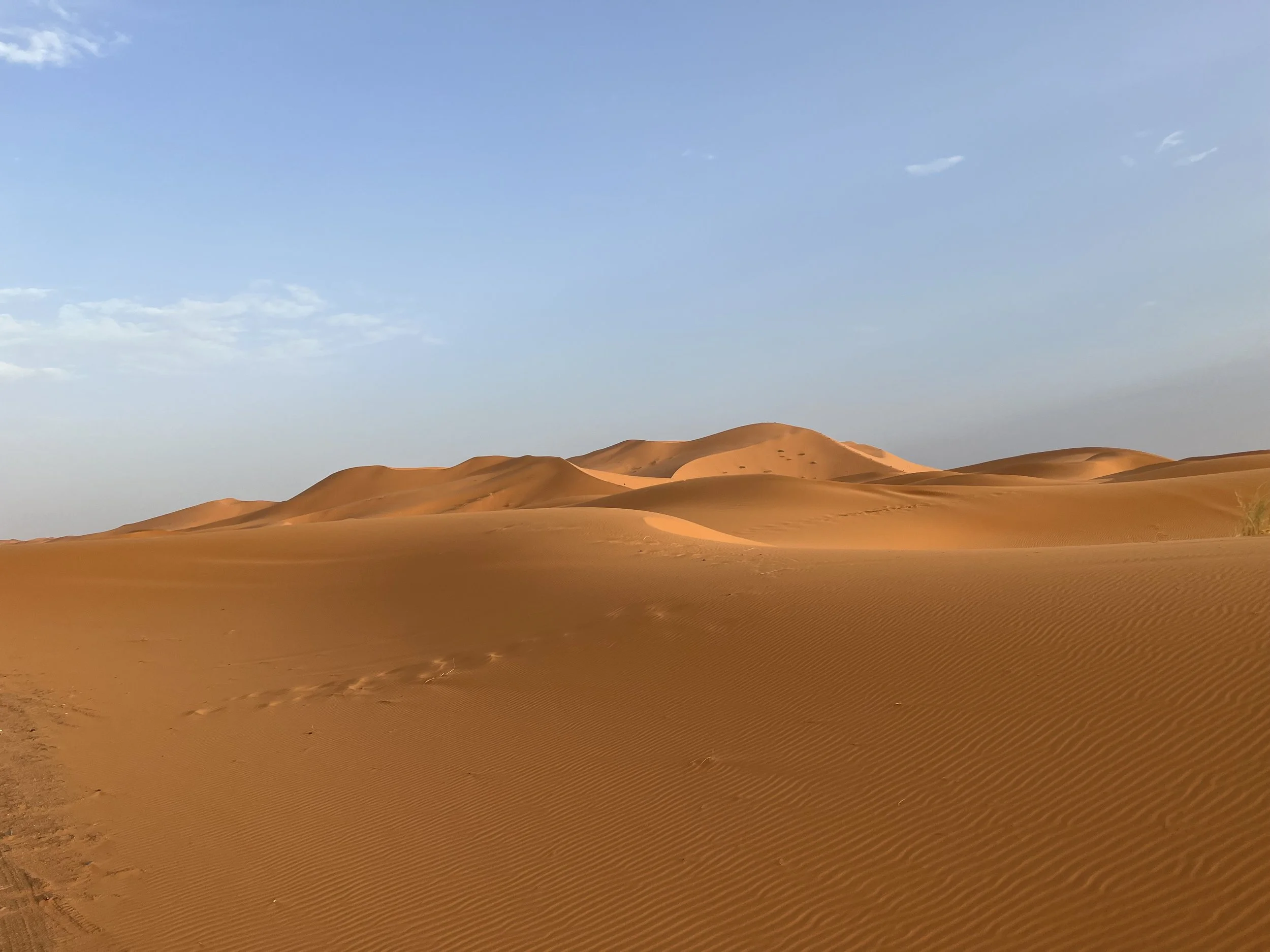 Photo of sand dunes in a desert with a clear blue sky and some clouds in the background.