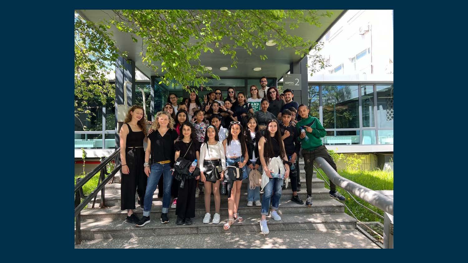 A group of diverse young people standing on steps outside a modern building with glass windows and a tree overhead.