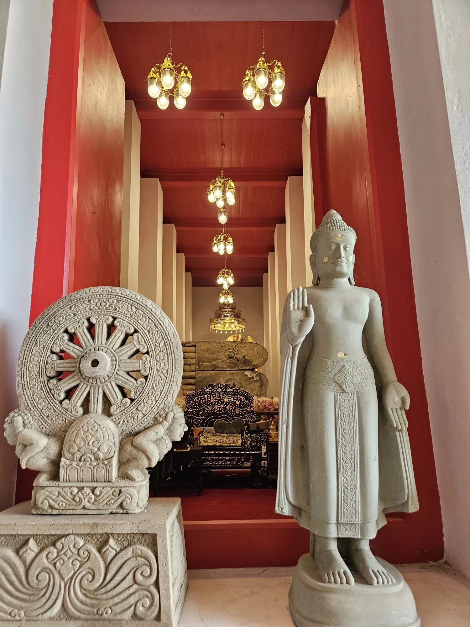 A white Buddha statue with an upright posture and hand raised in blessing stands beside a carved stone wheel and two lions on a pedestal, inside a room with red walls, a wooden ceiling, and hanging chandeliers.