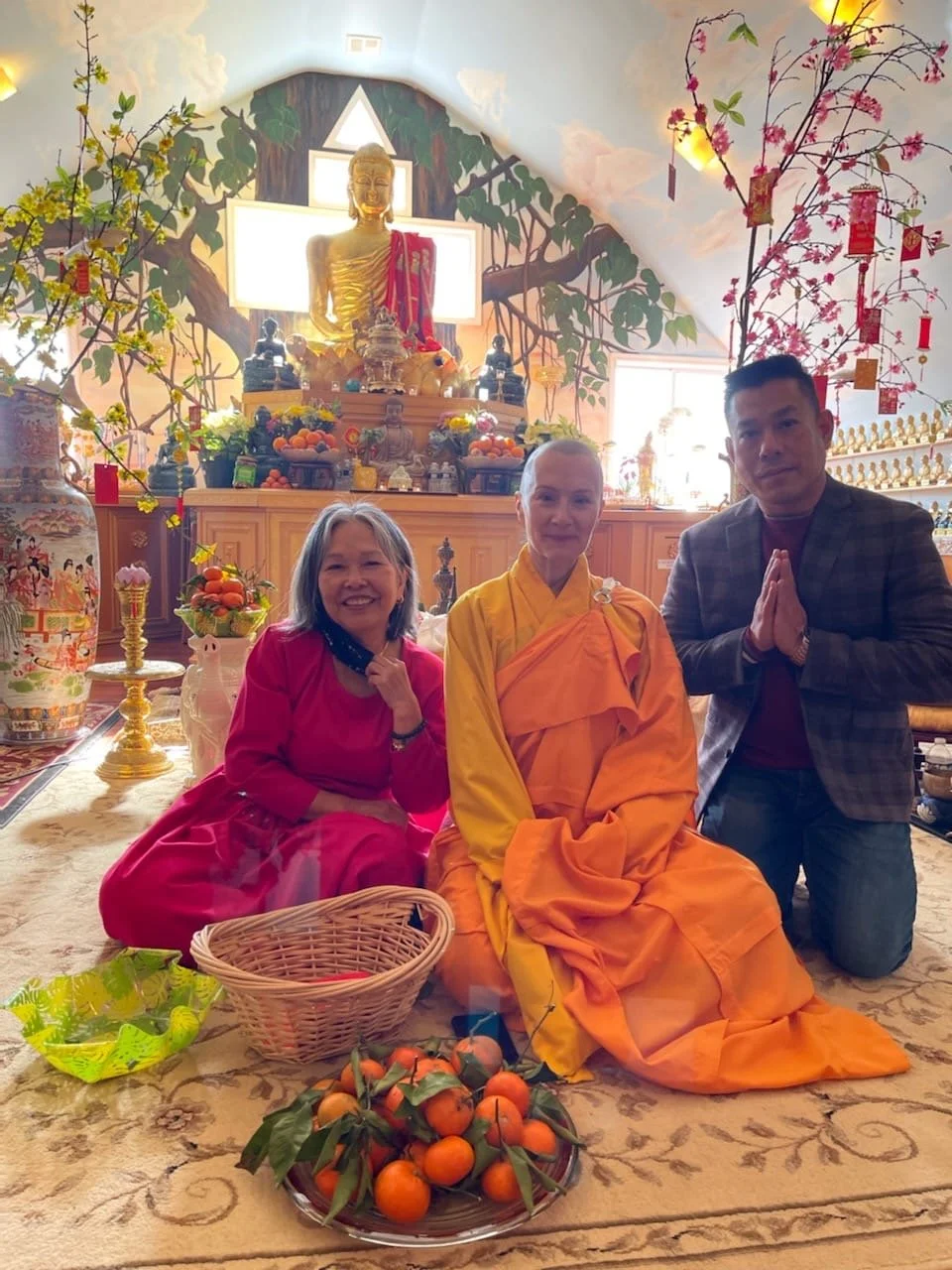 People kneeling on a rug in front of a Buddha statue during a cultural or religious ceremony, surrounded by offerings including oranges and flowers inside a decorated room.