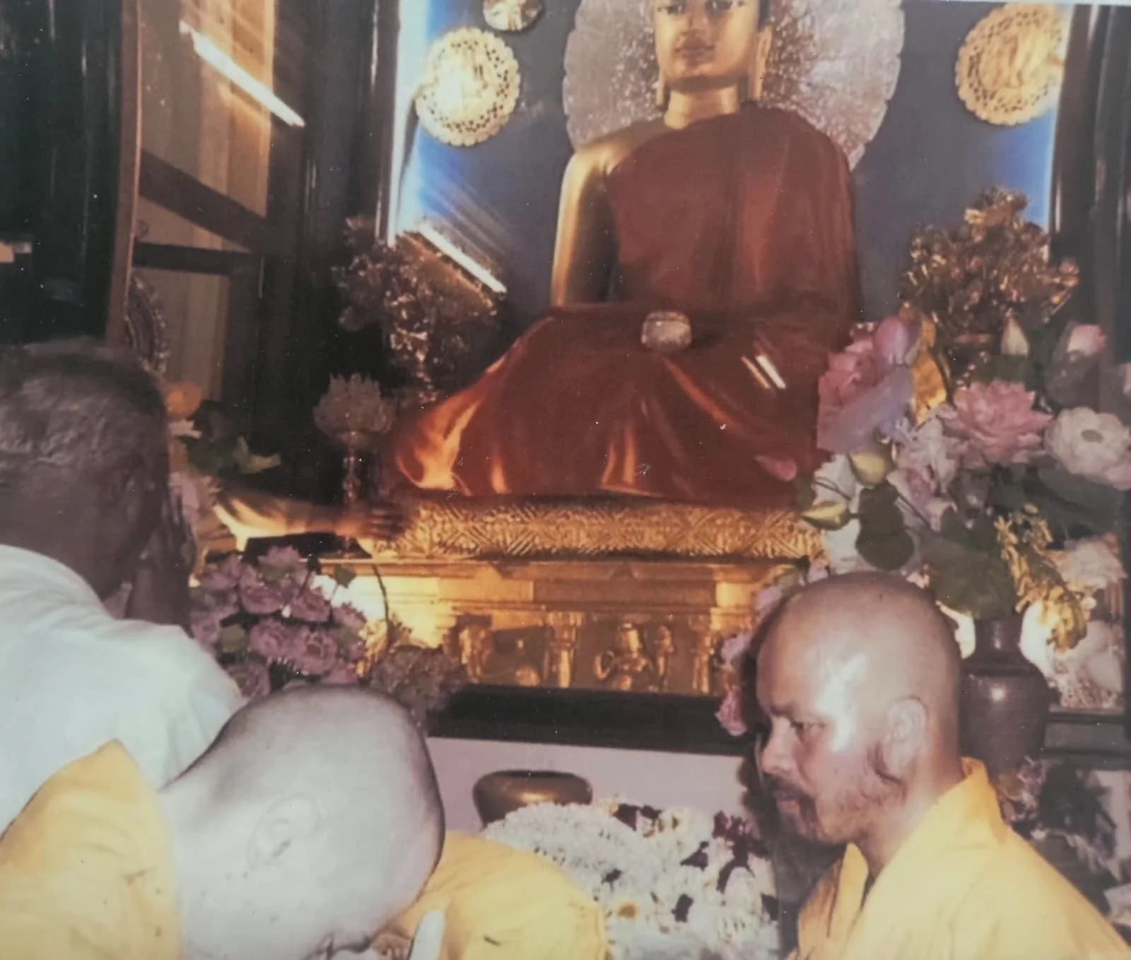 A group of monks in yellow robes participating in a religious ceremony in front of a large statue of Buddha. The Buddha statue is seated, draped in a reddish-brown cloth, with a serene expression, and is surrounded by flowers and offerings.