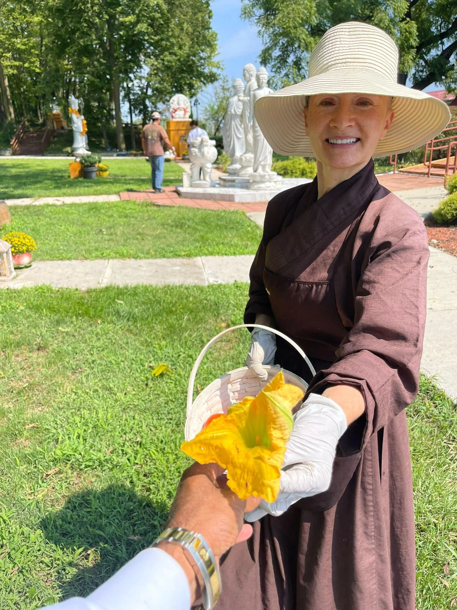 A smiling woman wearing a wide-brimmed hat and a brown robe extending her hand with a basket of yellow flowers. The scene is outdoors with greenery and statues in the background.