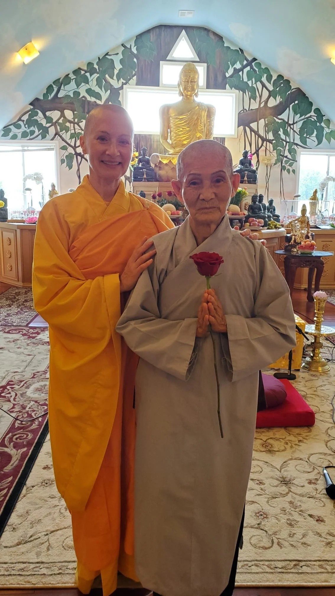 A monk in grey robes holding a red rose, standing next to a woman in yellow robes, inside a room with religious Buddhist statues, artwork, and decorations.