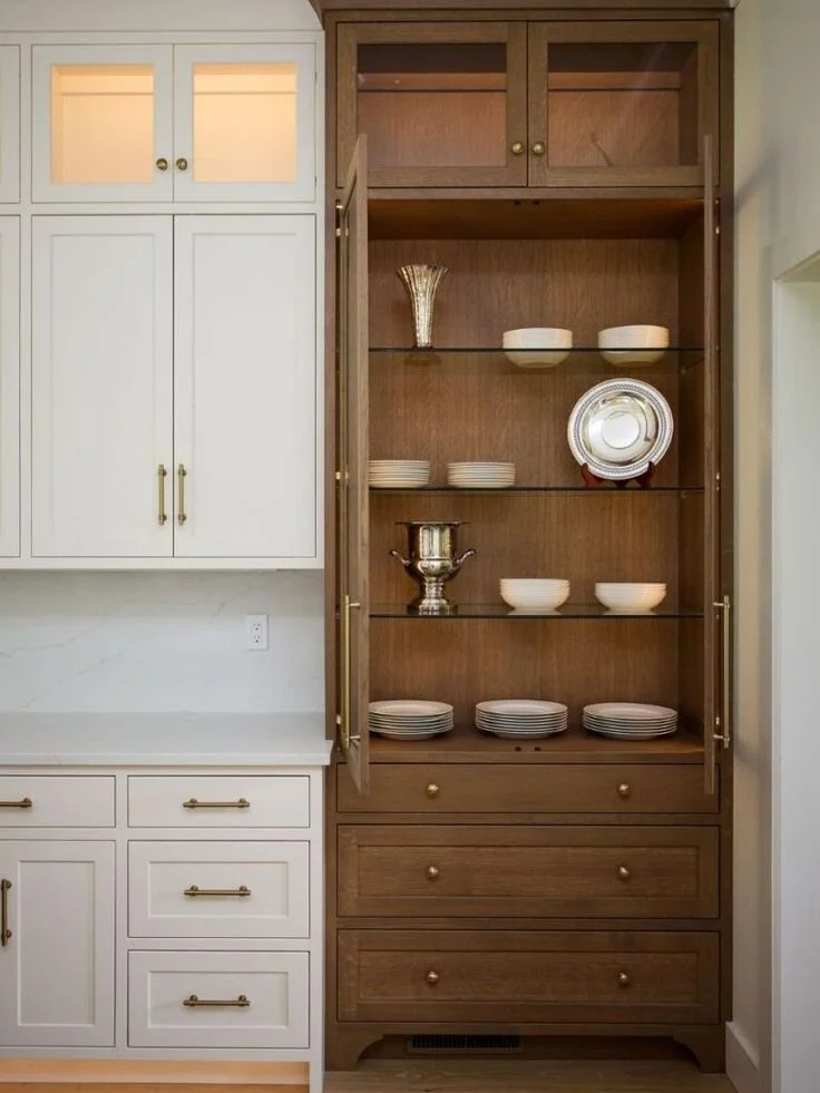 Two-tone kitchen cabinetry featuring white shaker cabinets paired with a warm wood glass-front hutch, brass hardware, and built-in storage, showcasing timeless custom cabinetry design.