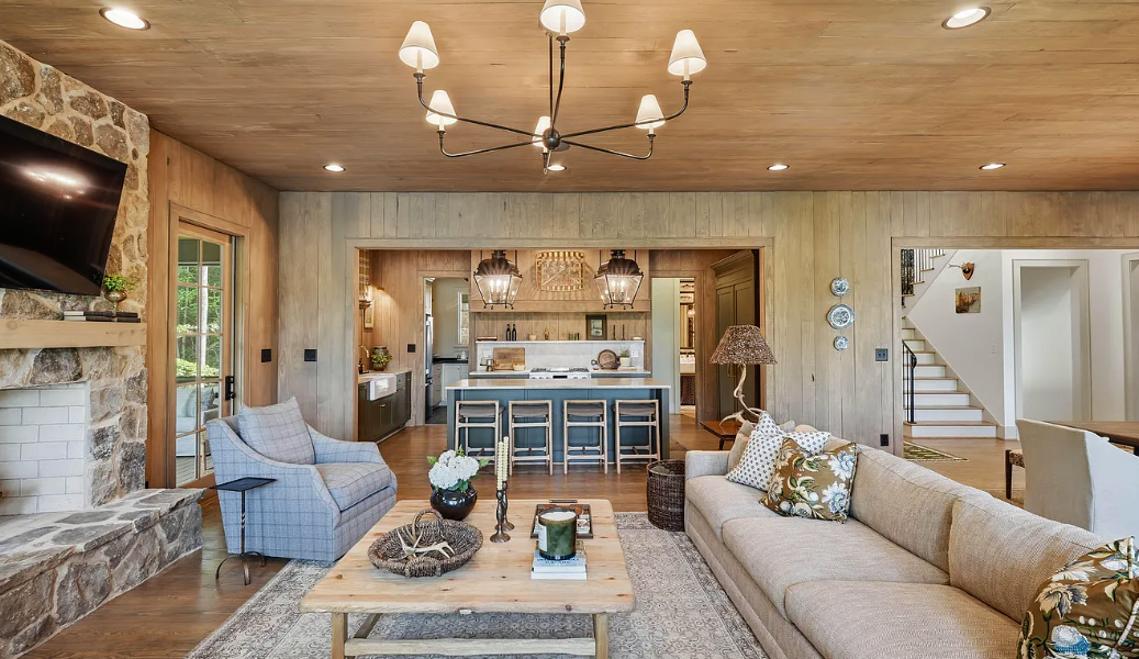 Collected living room with exposed wood beams, a stone fireplace, neutral seating, and an open kitchen beyond.