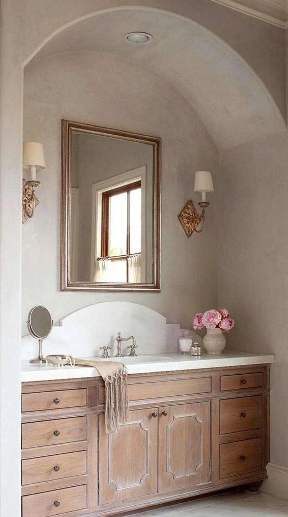 Traditional bathroom vanity with warm wood cabinetry, decorative door detailing, arched wall niche, and classic sconce lighting.
