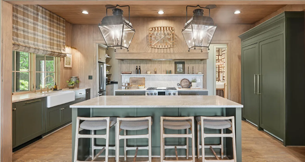 Green kitchen cabinetry with a pale island, four counter stools, lantern pendants, and a warm wood ceiling.