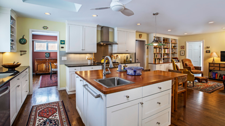 White kitchen cabinetry with a functional island, integrated storage, and open living layout designed by Angelini and Associates Architects, showcasing cabinetry as a central design anchor in a residential renovation.