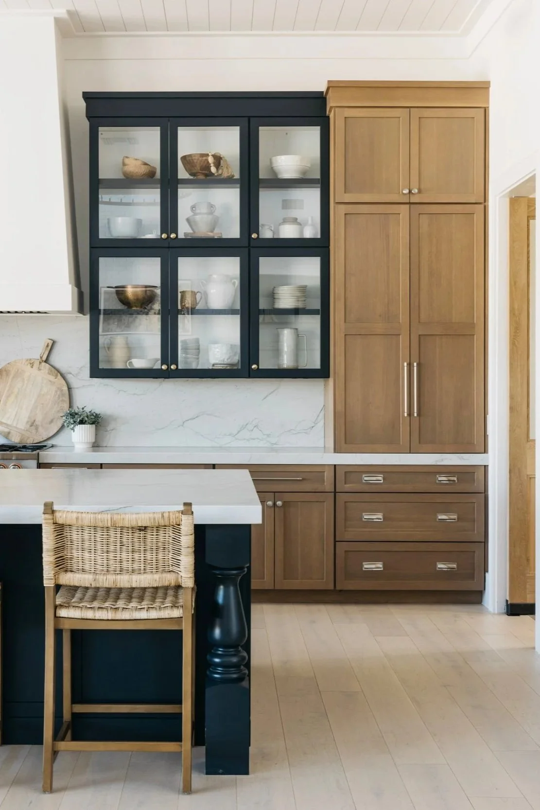 Kitchen cabinetry featuring dark glass-front upper cabinets, warm wood pantry cabinets, brass hardware, and light wood flooring in a bright, neutral kitchen.