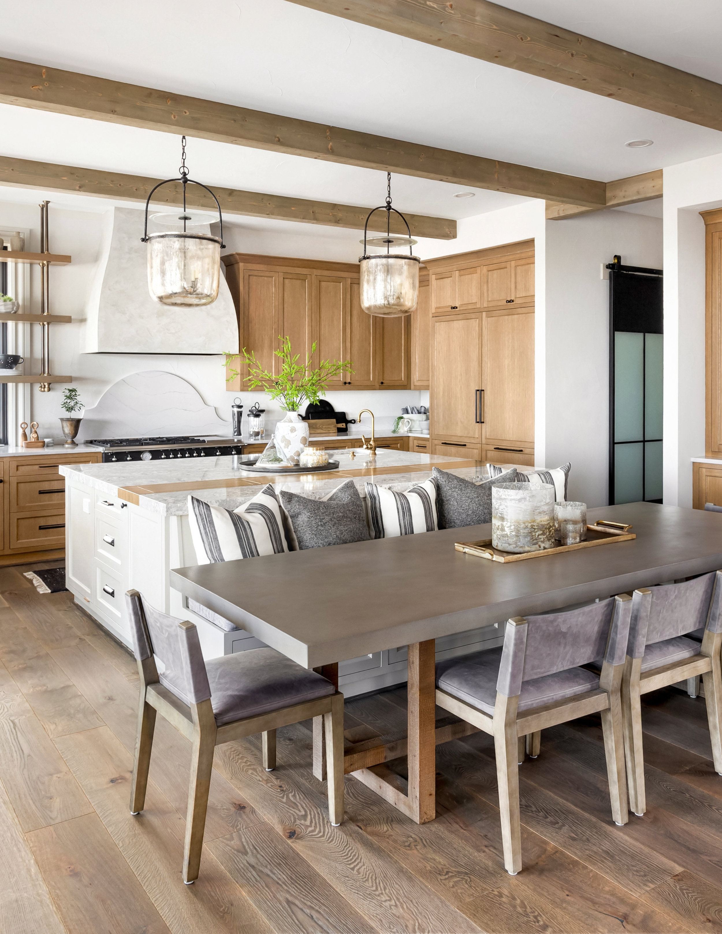 Two-tone kitchen featuring natural wood cabinets, white island cabinetry, exposed wood ceiling beams, oversized glass pendant lights, and a dining table integrated into the kitchen layout.