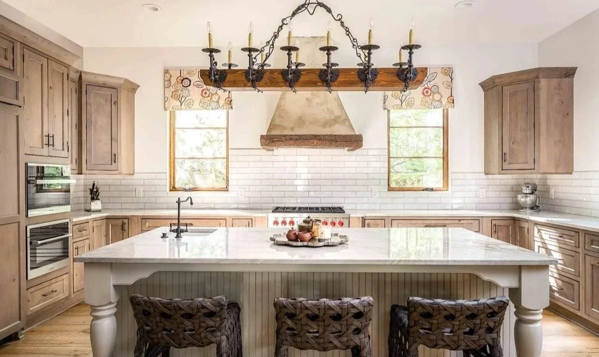 Calm traditional kitchen with a large stone island, woven stools, neutral cabinets, and a sculpted range hood.