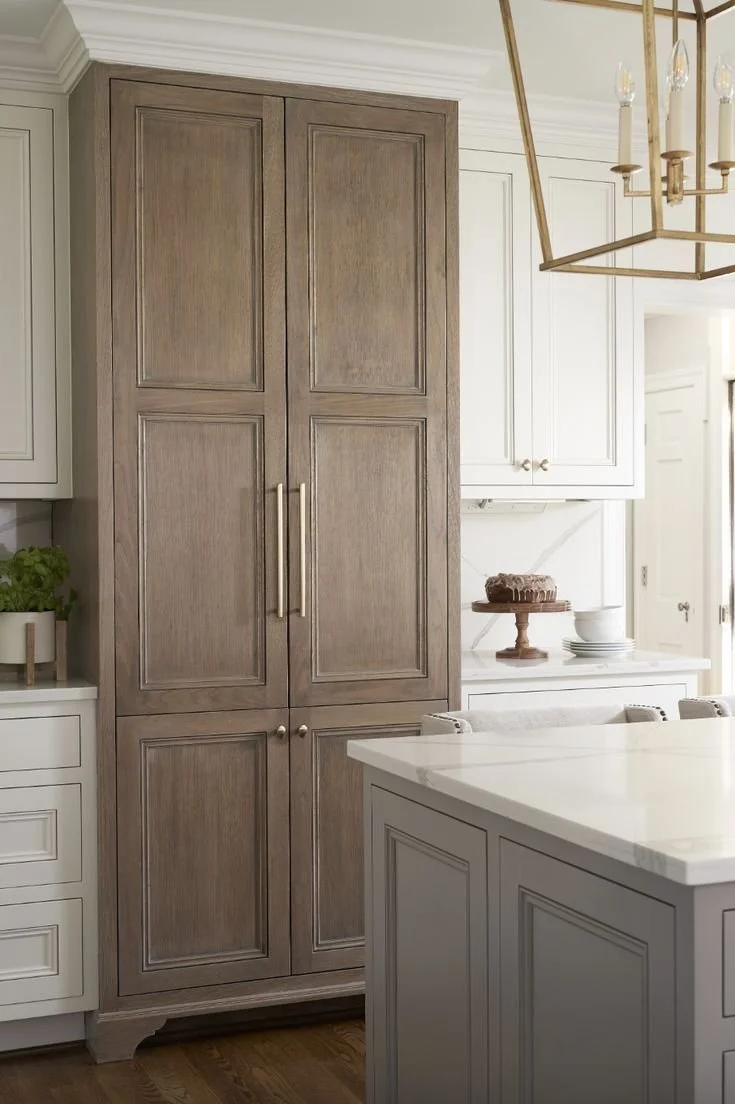 Tall warm wood pantry cabinets paired with soft painted kitchen cabinetry, showcasing a layered cabinet design with natural materials and timeless proportions.