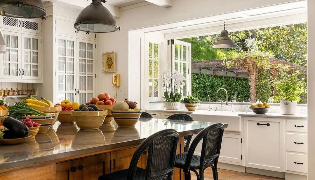 Bright white kitchen with glass-front cabinets, dark countertops, a wood island table, and a garden window.