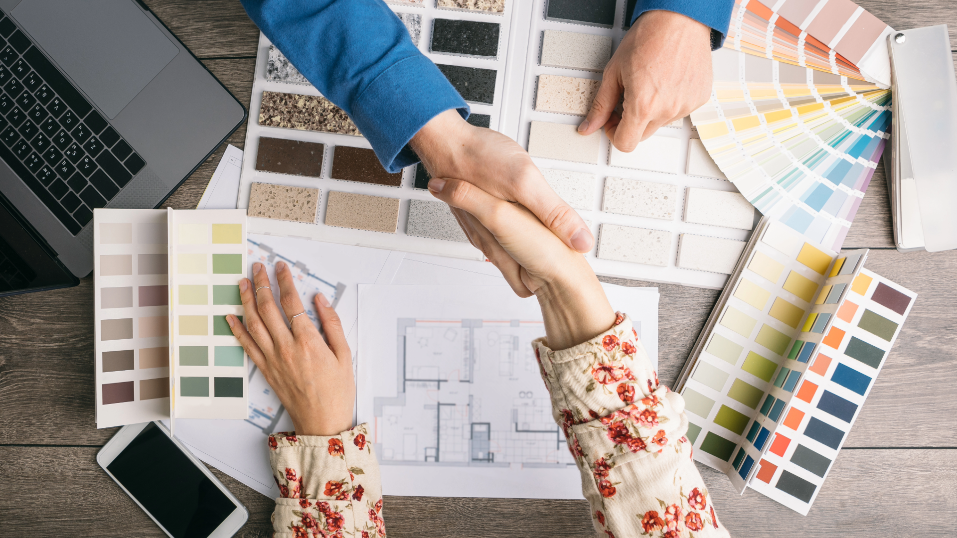 Homeowner and cabinetry designer shaking hands over kitchen design plans with countertop samples and color palettes.