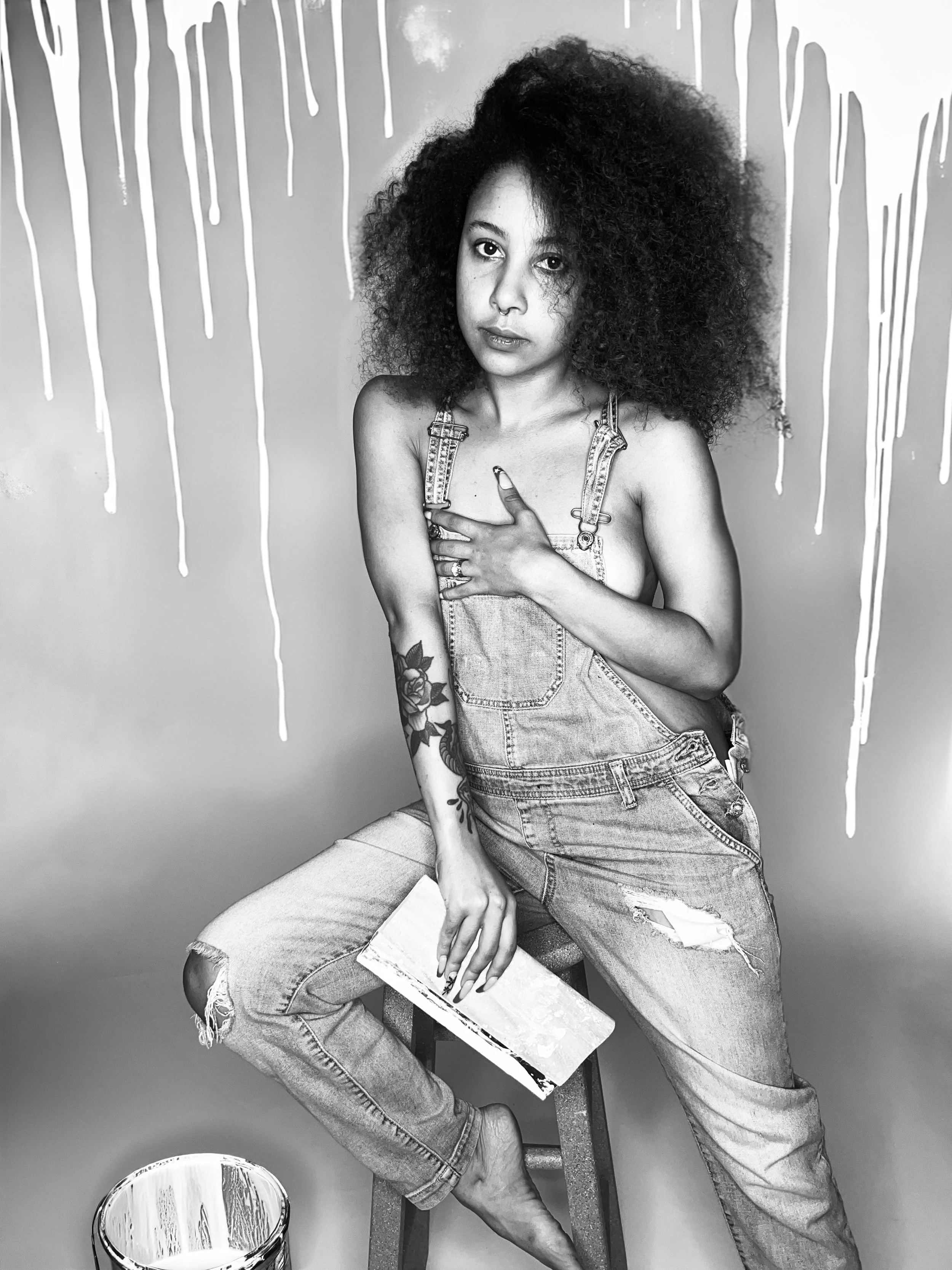 Black and white photograph of a person with curly hair sitting on a stool. They are wearing denim overalls with ripped knees, holding a large paintbrush. White paint drips on the wall behind them, with a paint can on the floor.