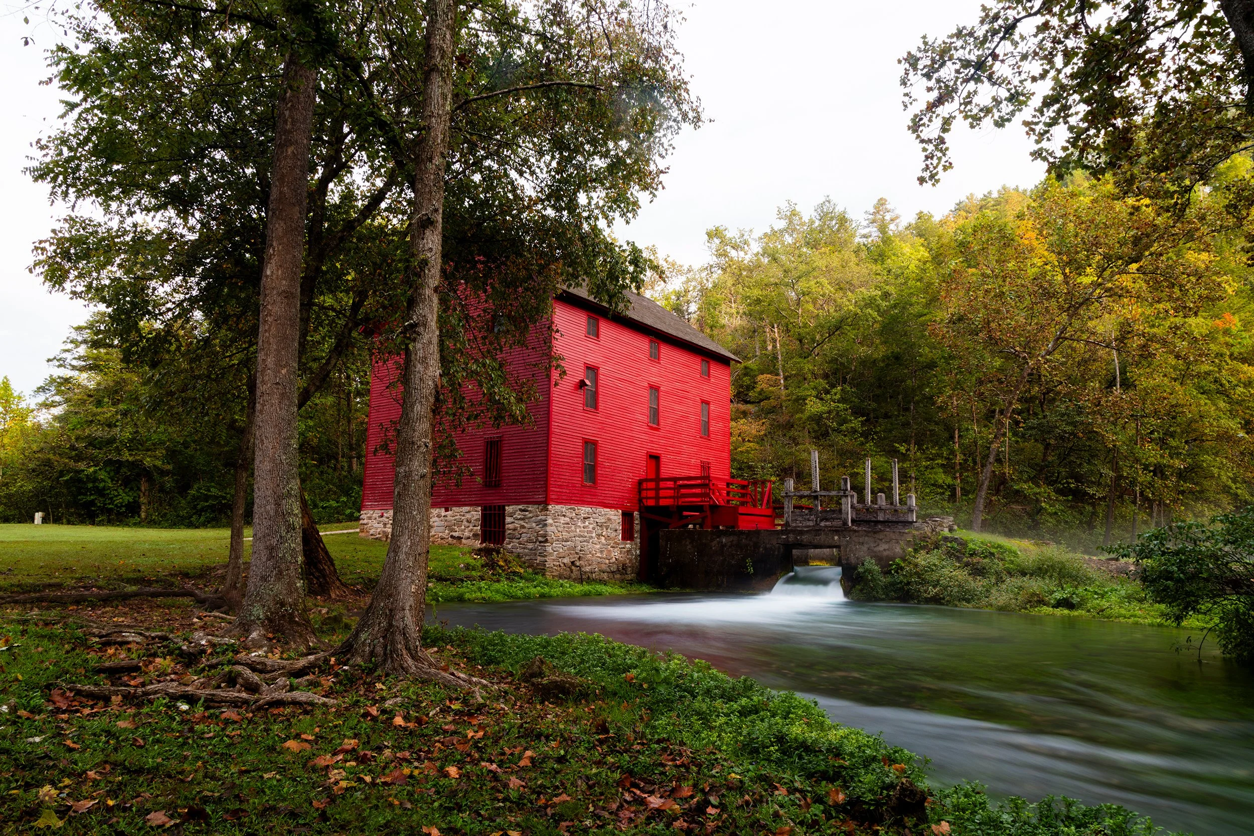 A red historic mill house beside a flowing river surrounded by green trees.