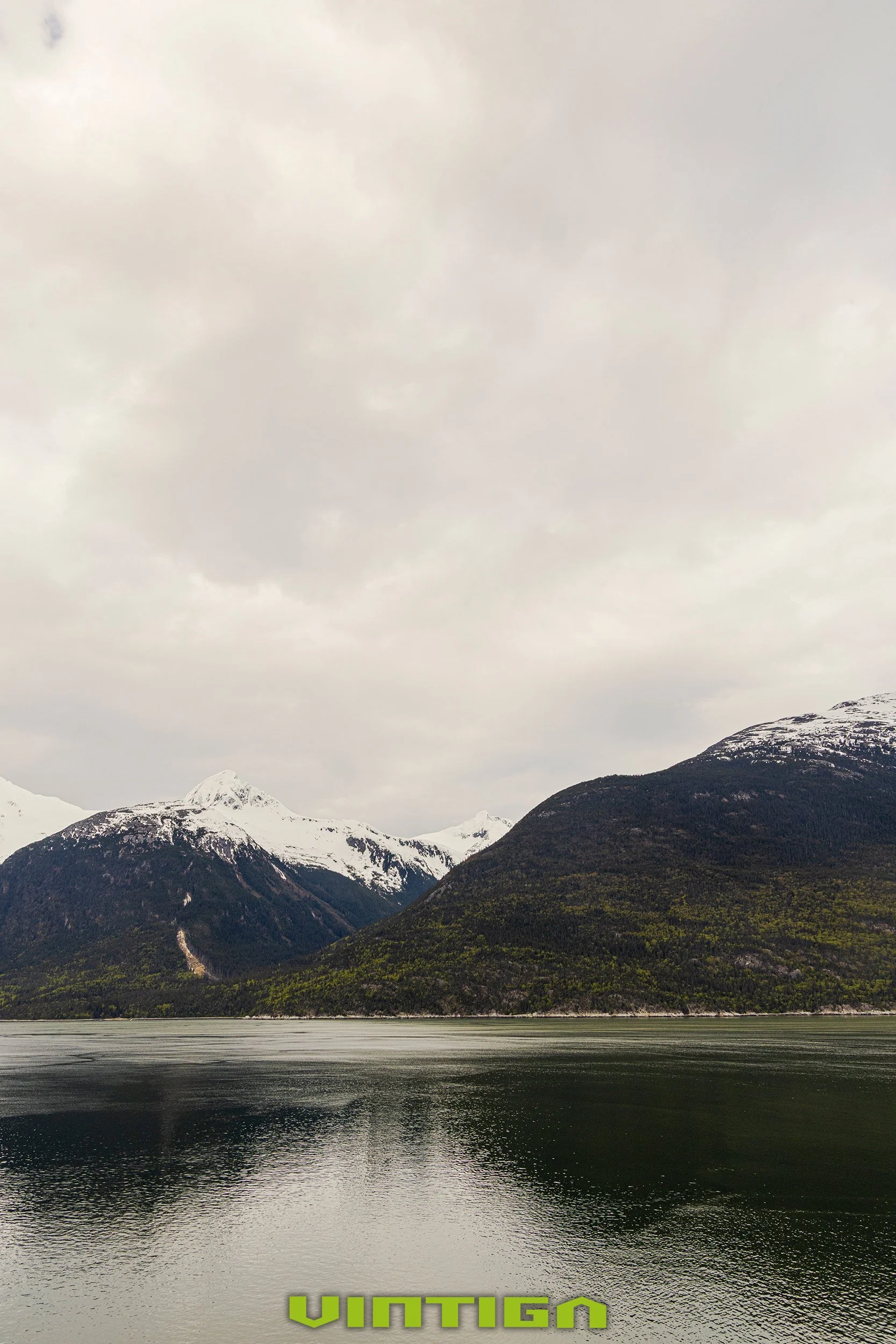 A mountainous landscape with snow-capped peaks, green slopes, and a body of water in the foreground under an overcast sky.