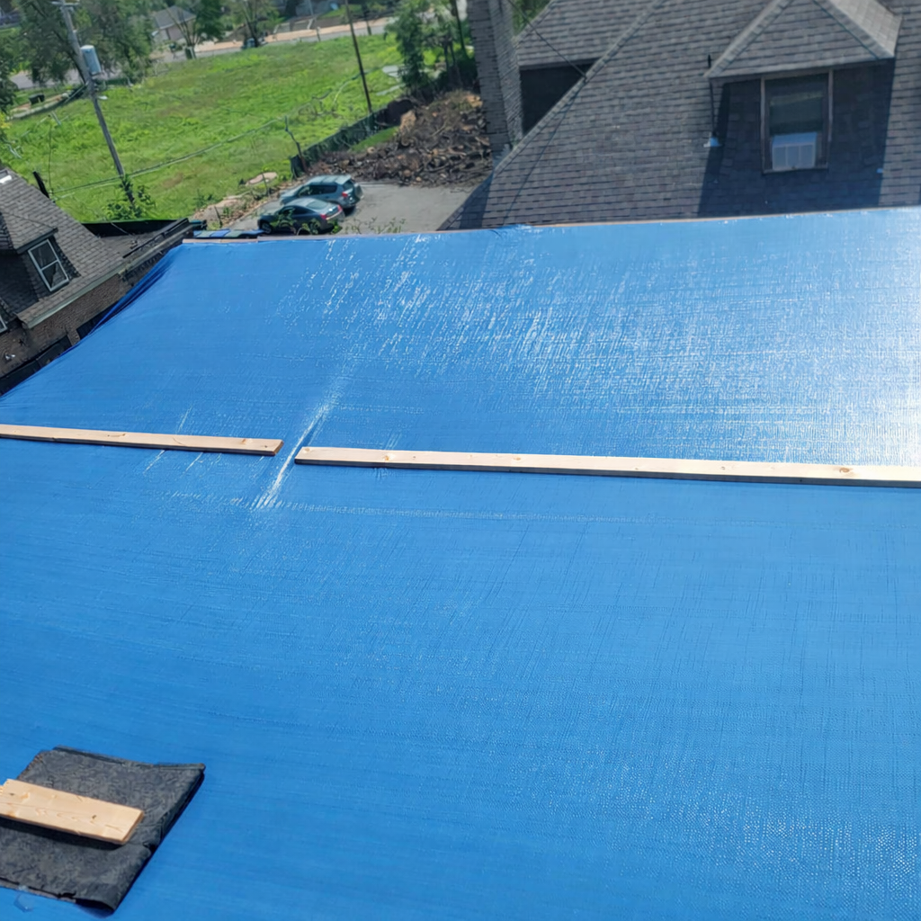 A blue tarp is stretched over a rooftop, secured with wooden strips, with a house roof and some greenery visible in the background.