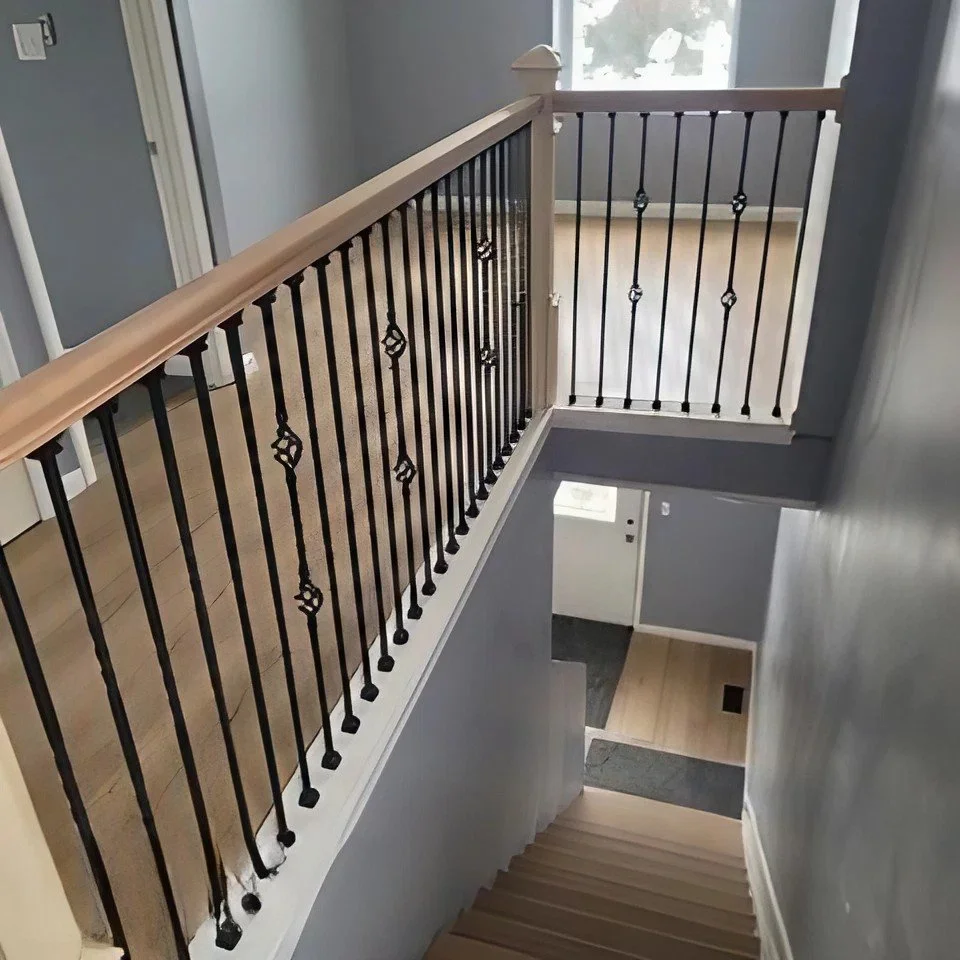 Interior view of a staircase with black wrought iron railing and light wood handrail, looking down from upstairs to the ground floor, with a window and door in the background.