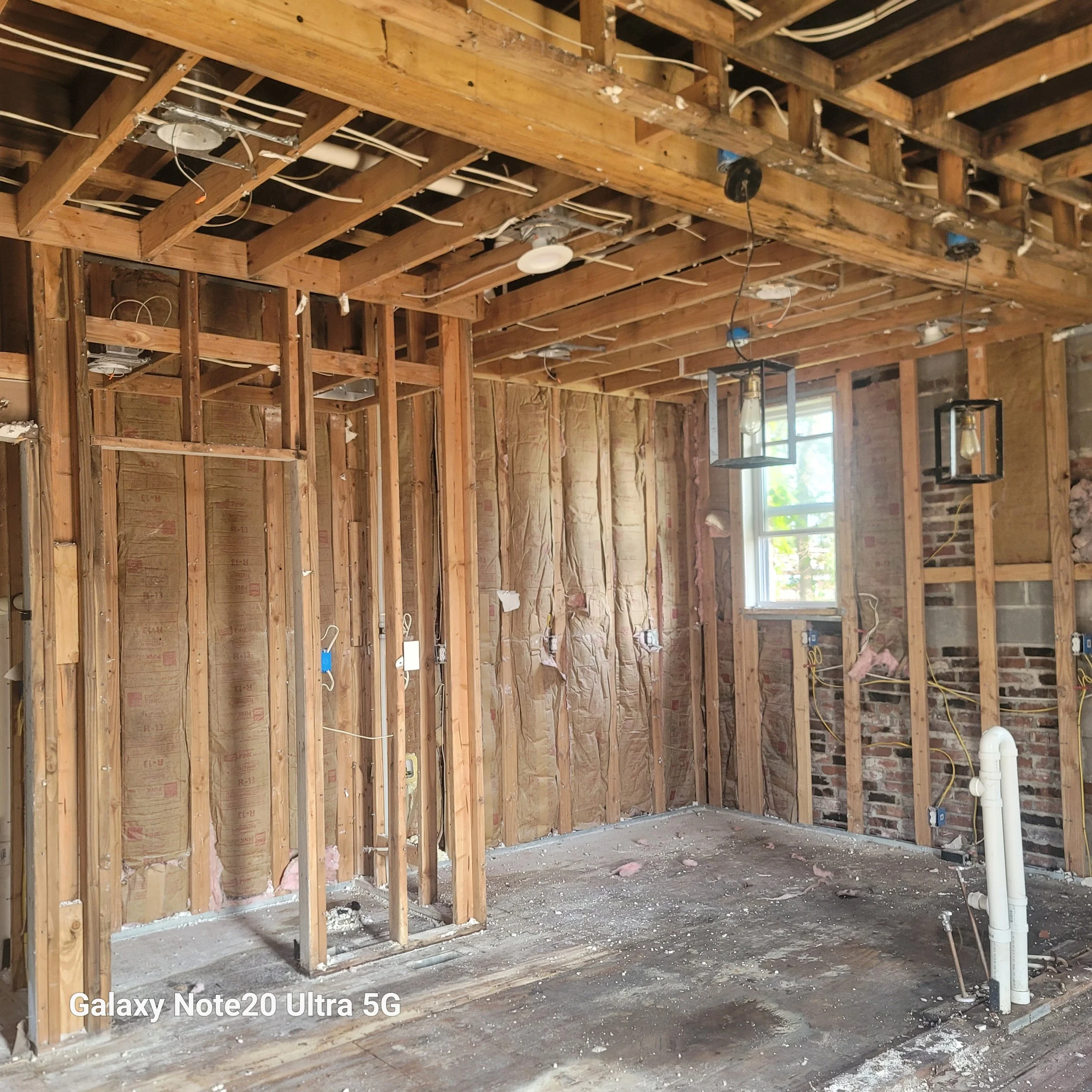 Interior of a house under renovation with exposed wooden studs, insulation, and electrical wiring. There is a window on the wall, and hanging light fixtures without bulbs.