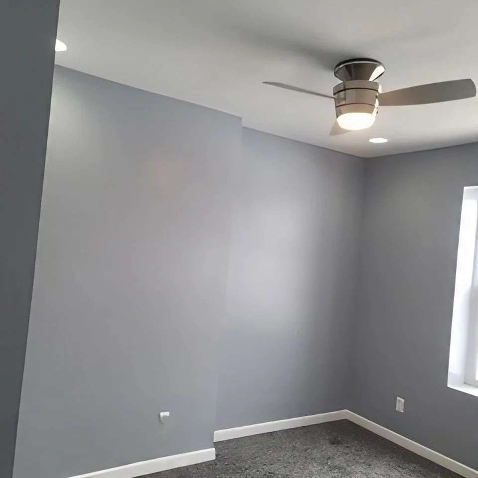 Empty room with gray walls, white baseboards, dark gray carpet, window with white trim, and ceiling fan with light.