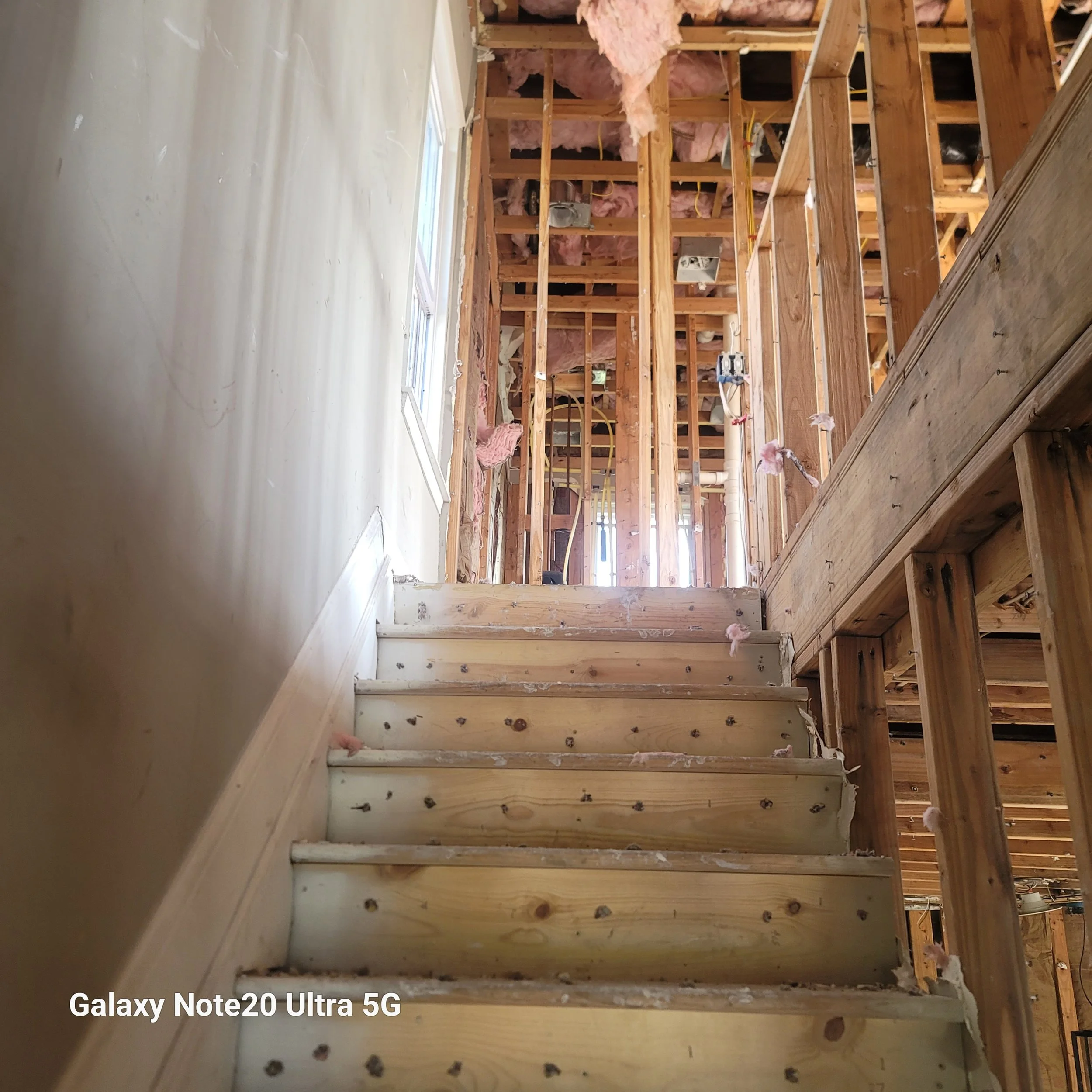Interior view of a house under construction showing wooden framing, insulation, wiring, and a stairway leading to the upper floor.