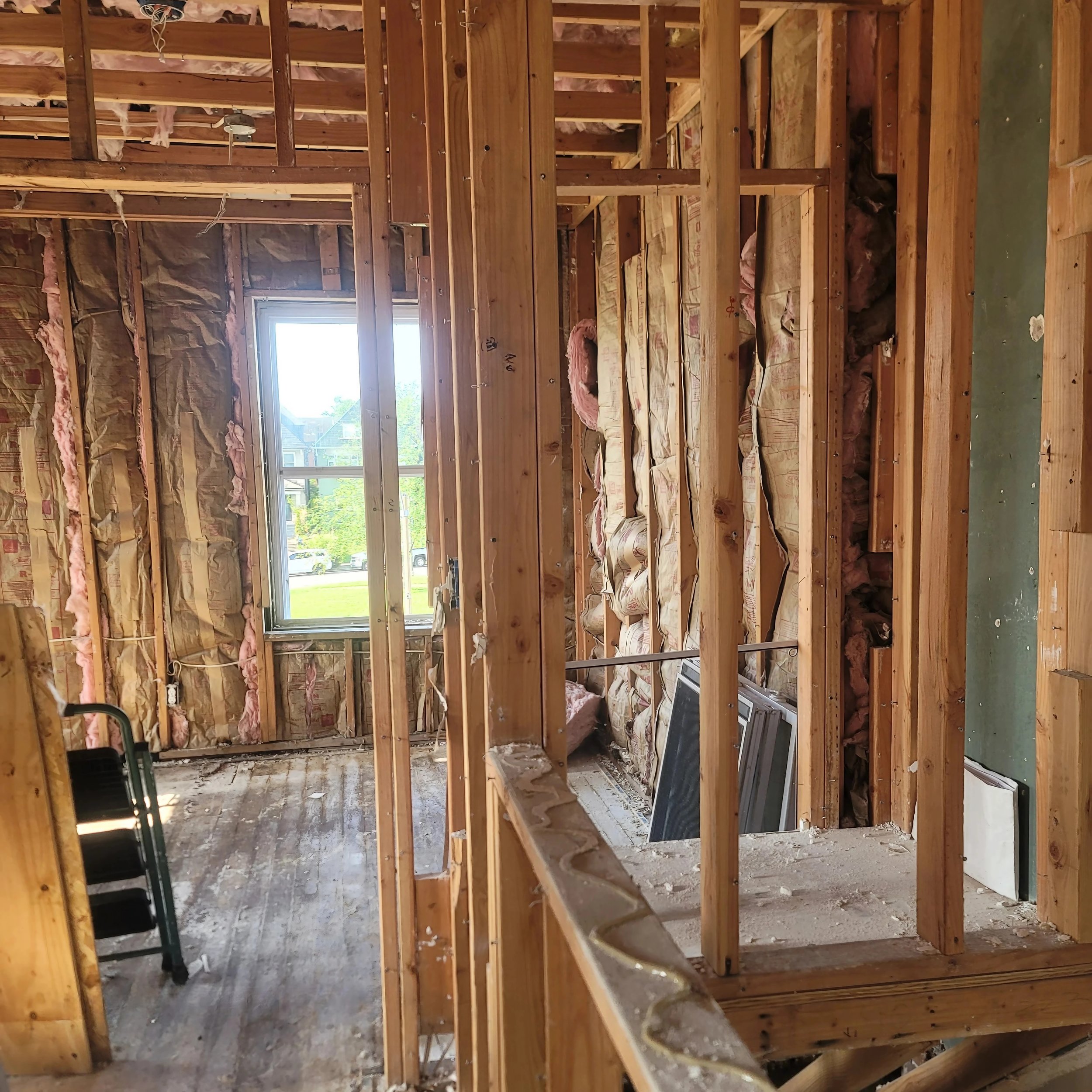 Interior view of a house under renovation, showing exposed wooden framing, insulation, and a window with a view of a grassy yard and houses outside.