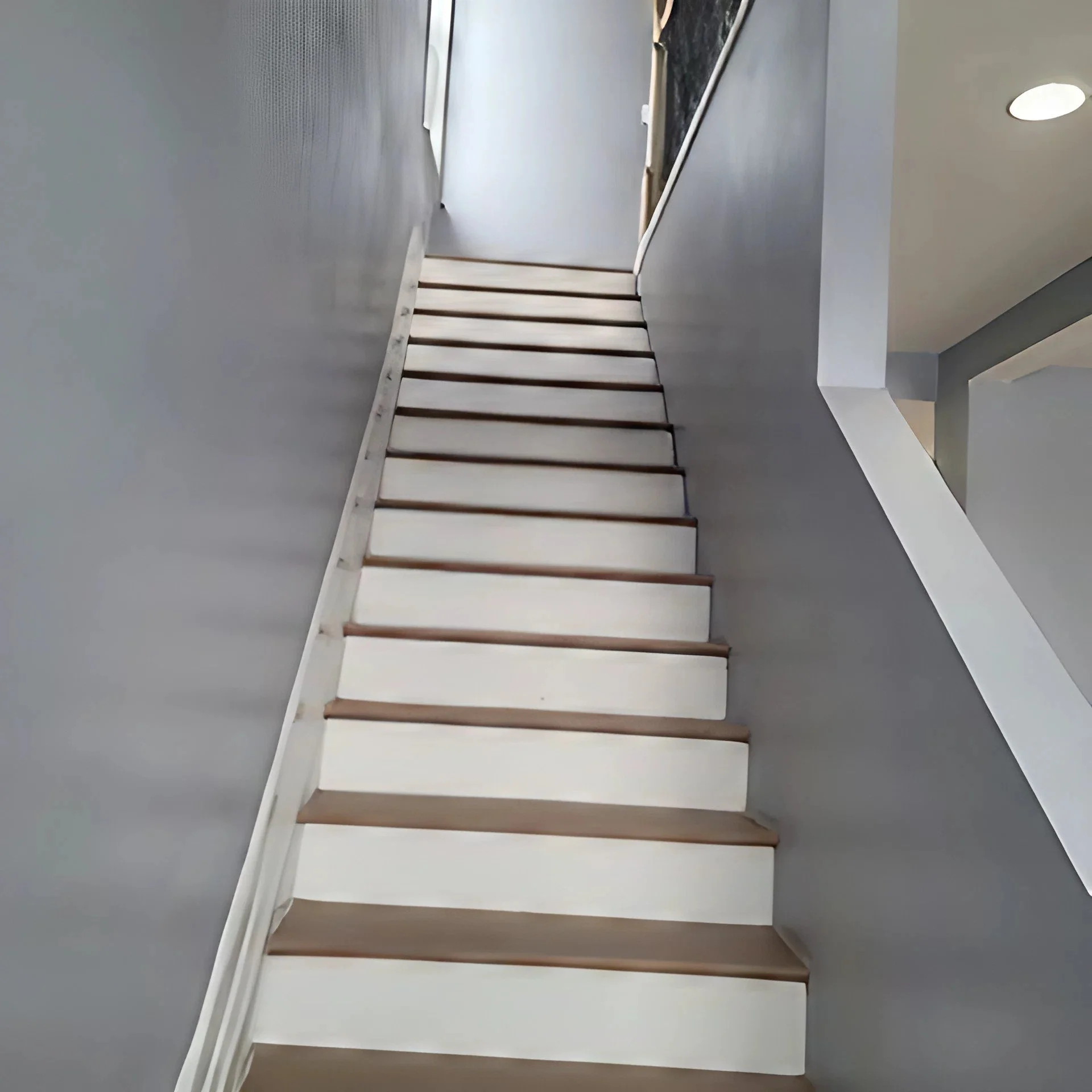 Interior view of a staircase with white risers and wooden treads, leading up to a hallway with gray walls and ceiling, with furniture and a window visible at the top.