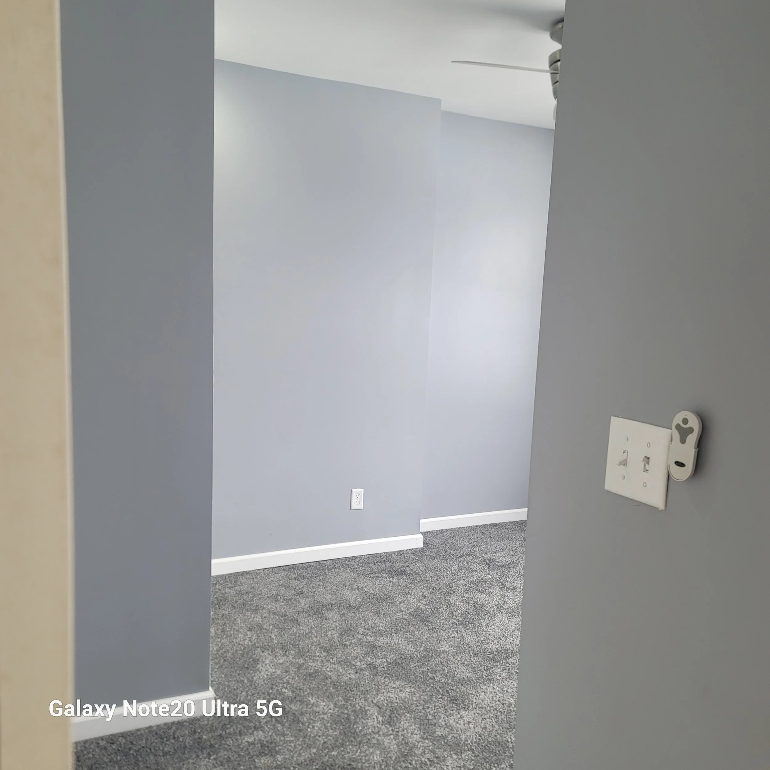 Empty room with light gray walls, white electrical outlet, and gray carpet flooring.