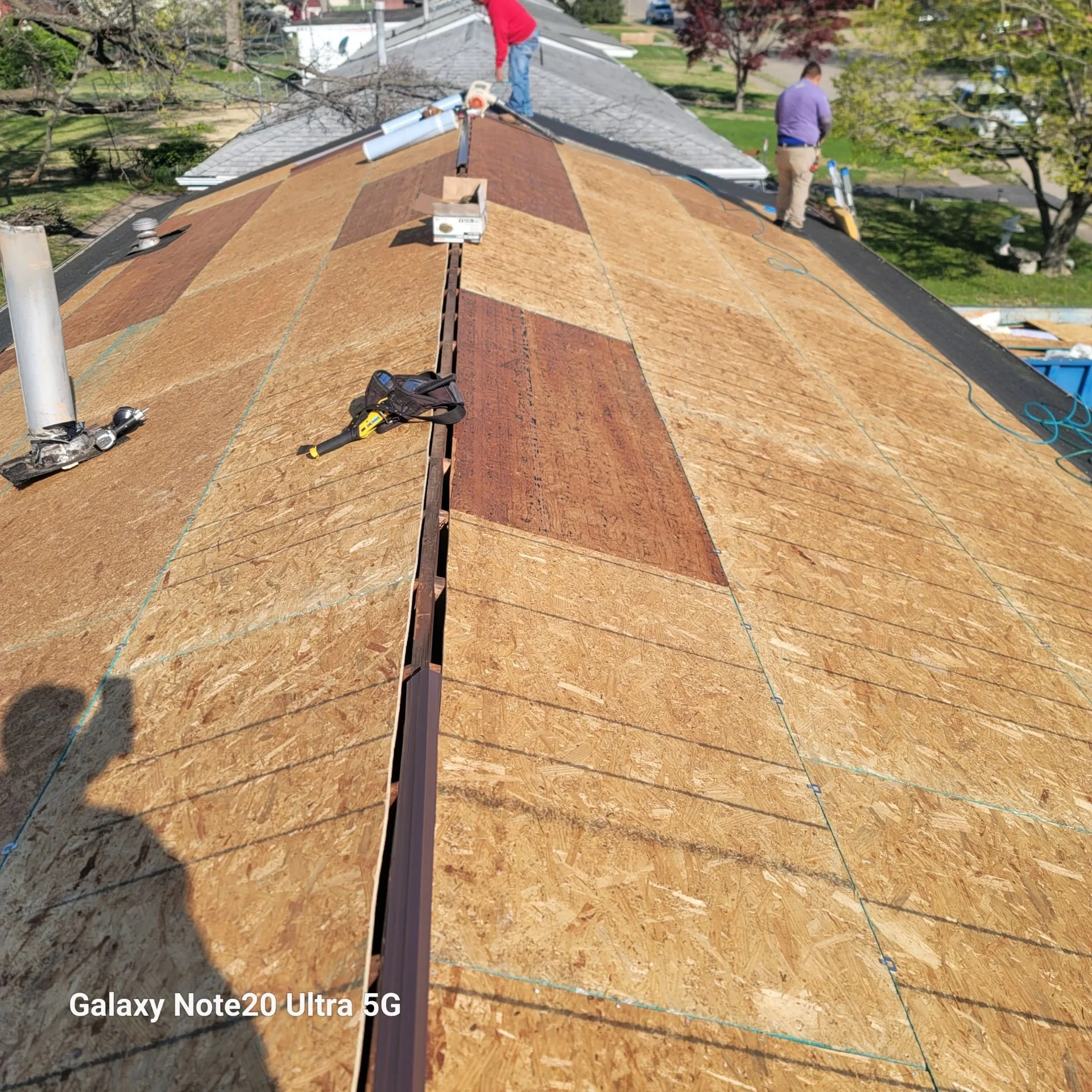 Workers installing new roofing on a house, with plywood sheets covering the roof and tools scattered around.