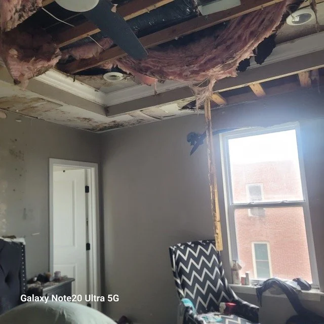 Room under renovation with exposed ceiling insulation, missing drywall, a window, and a patterned chair.