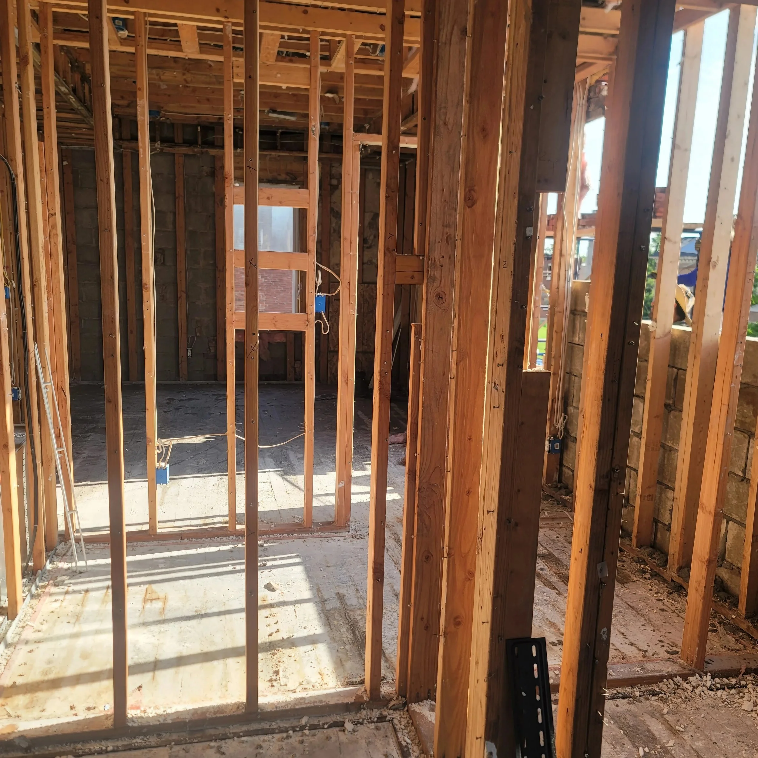 Interior view of a house under construction with exposed wooden framing, including upright studs, partial wall framing, and visible wiring, with sunlight coming through planned windows.