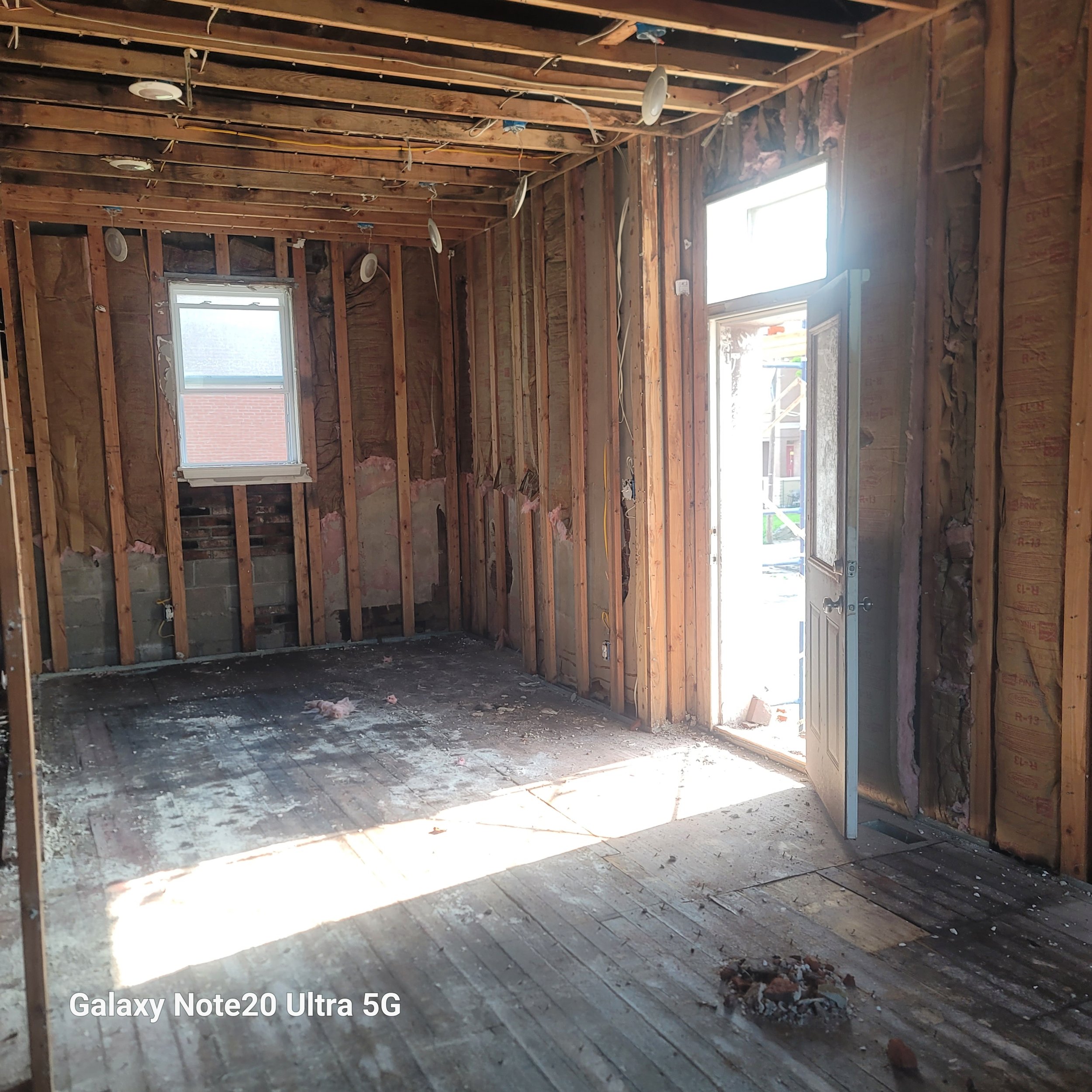 Interior of a room under construction with exposed wooden studs and framing, a small window, and an open door leading outside, with sunlight illuminating the unfinished floor.