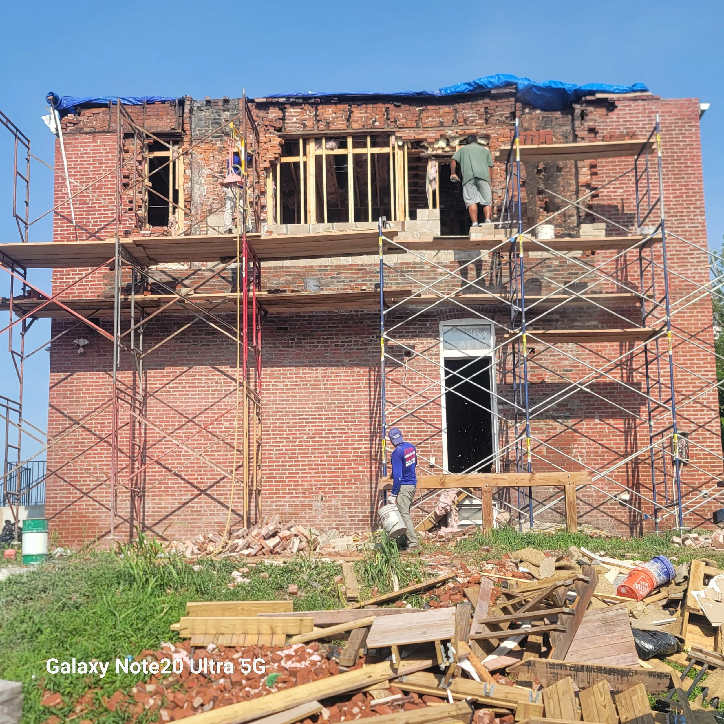 Construction workers rebuild a brick house with scaffolding and wood framing, debris scattered on the ground, under clear blue sky.