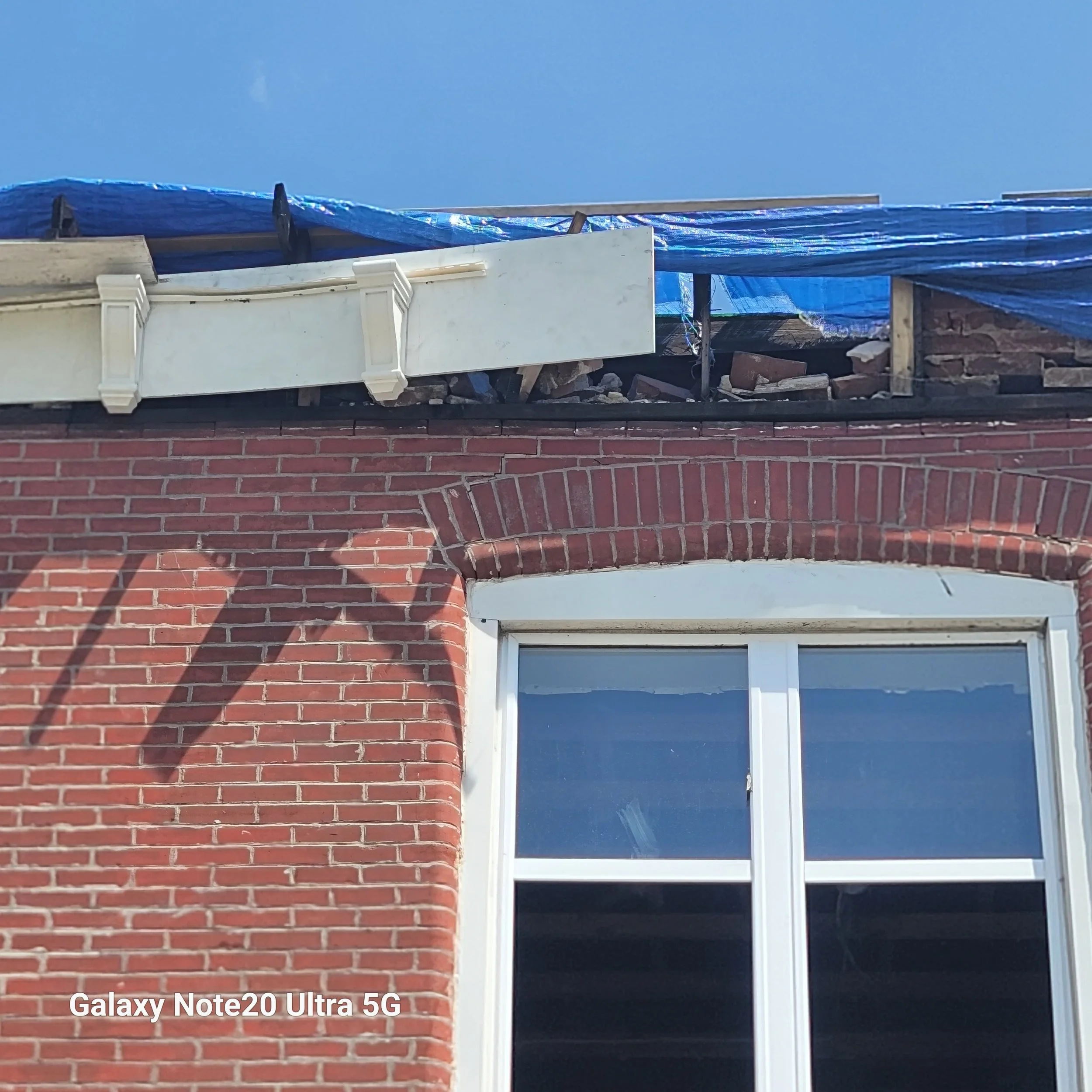 Brick building with a large window, and damaged roof with construction debris and blue tarp.