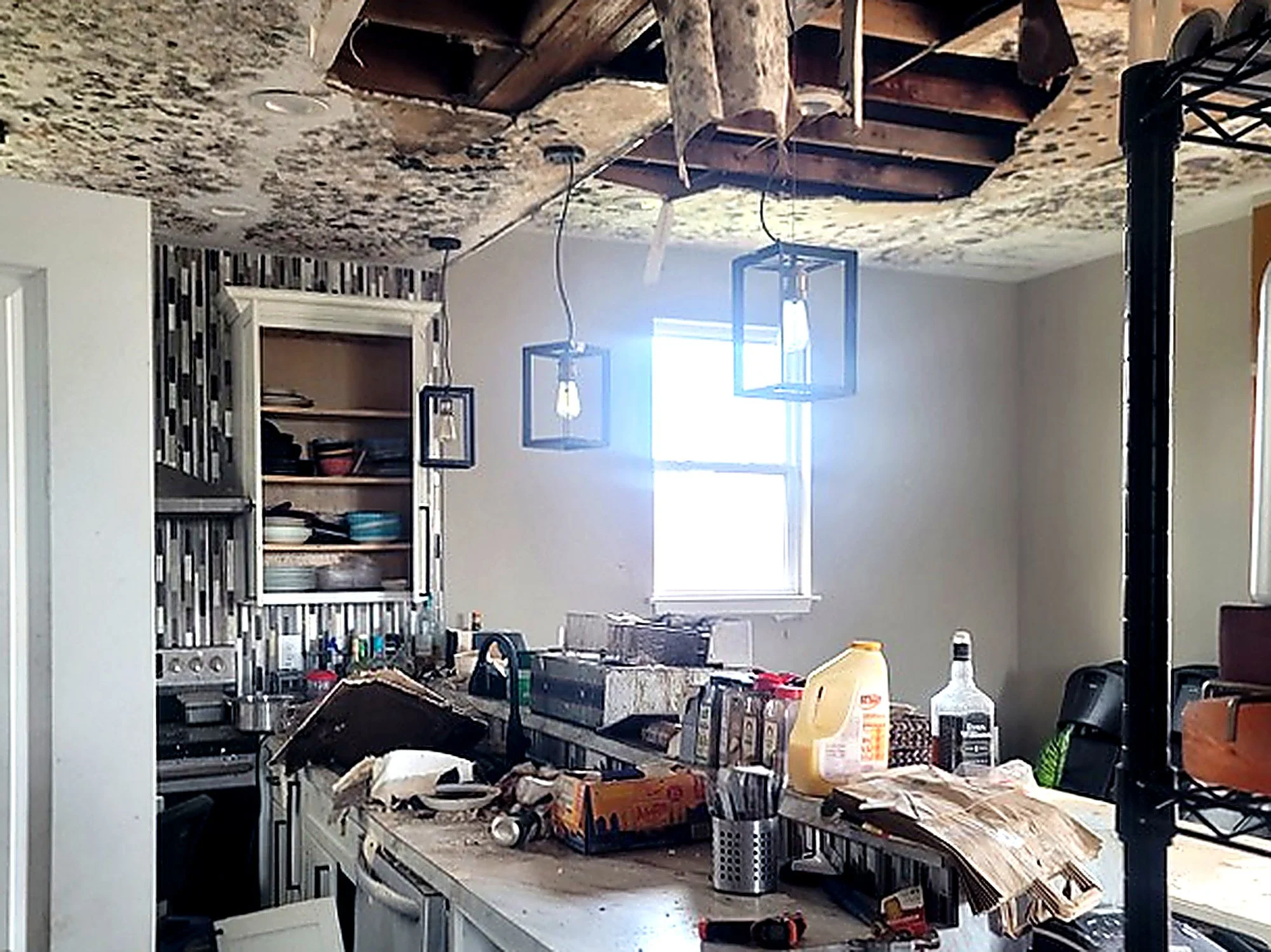 Kitchen under renovation with exposed ceiling, hanging light fixtures, cluttered countertop, open cabinets, and construction debris.