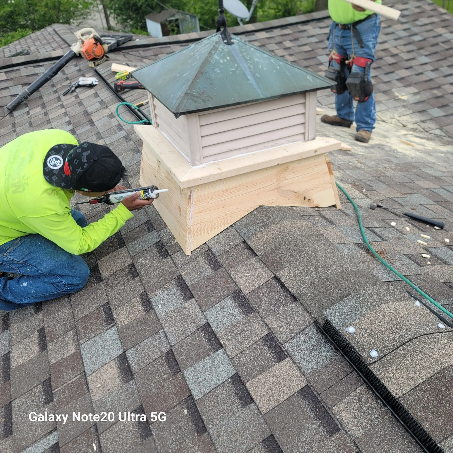 Workers installing or repairing a small roof vent on a residential roof, with one person applying sealant and another standing nearby.