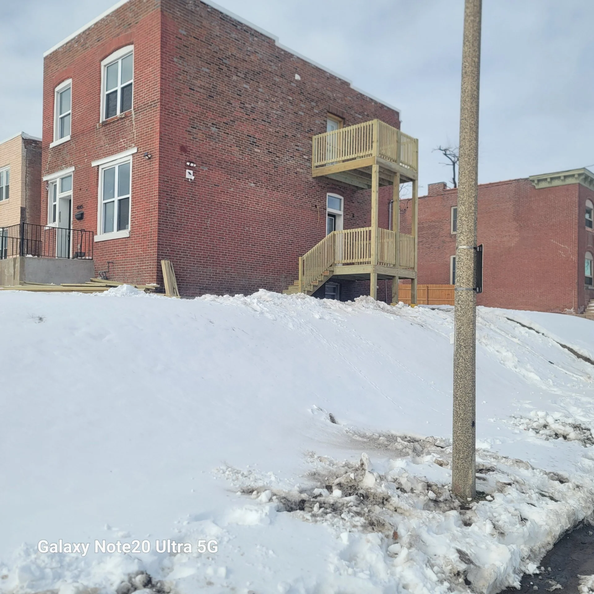 A multi-story brick building with large windows, a staircase leading to a balcony, and snow-covered ground with a pole in the foreground.