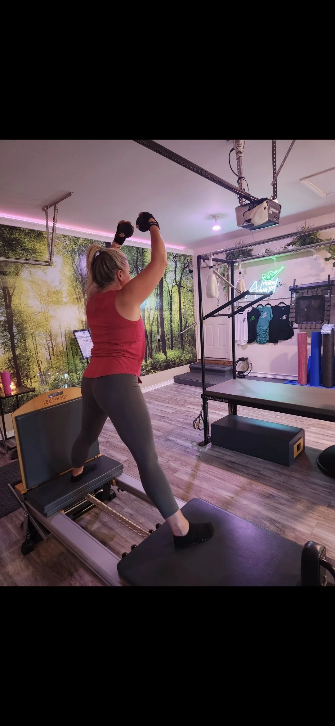 A woman in athletic clothing is exercising on a pilates reformer machine in a fitness studio decorated with a forest mural background and neon signs.