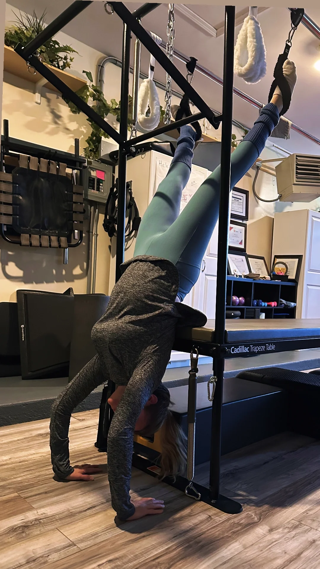 A person performing a headstand in a gym, with feet elevated and supported by a frame. The person is wearing leggings, a long-sleeve shirt, and socks.
